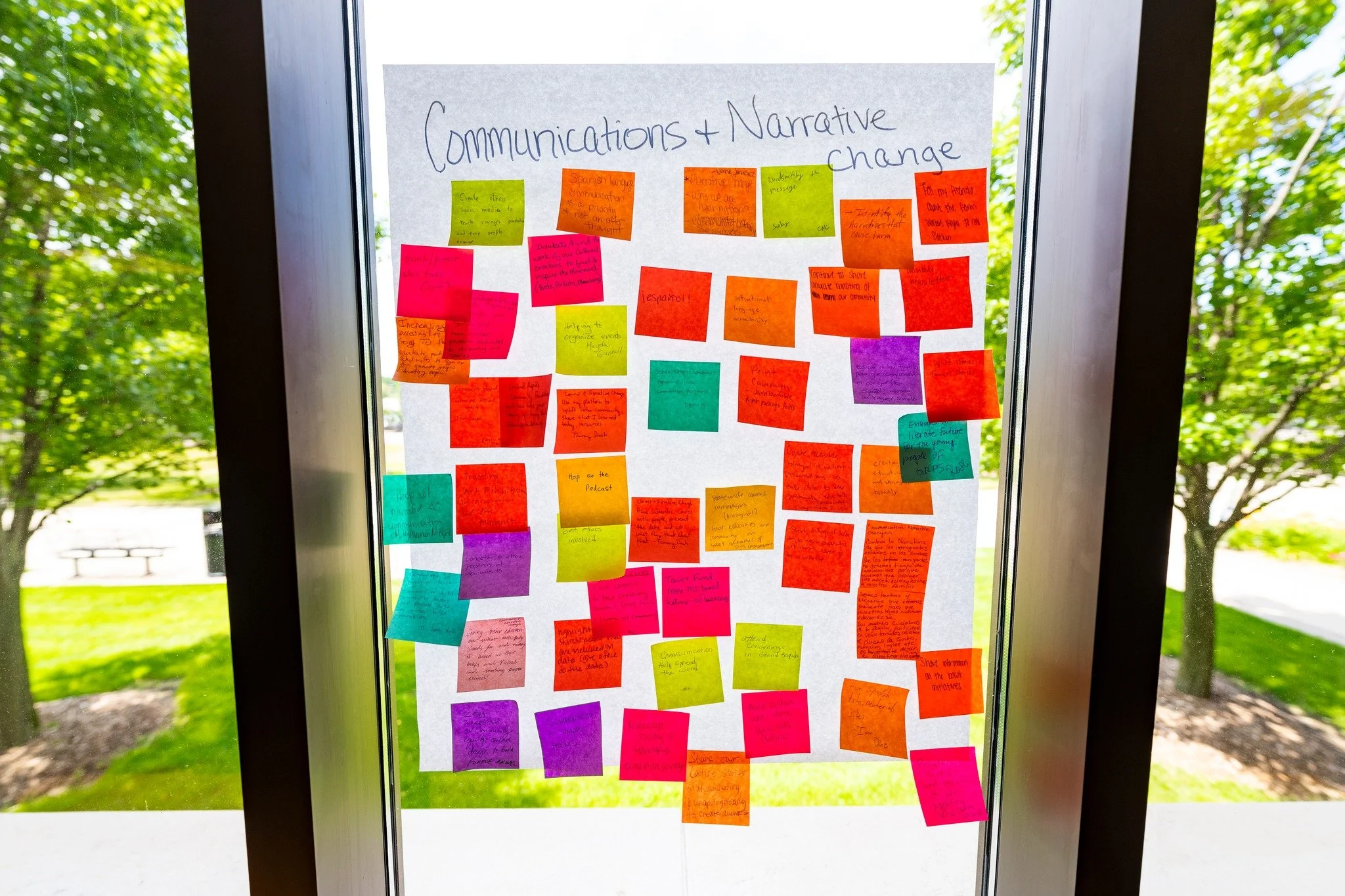 A poster on a glass door with colorful sticky notes about 'Communications + Narrative Change'. The poster is outside with green trees and grass visible in the background.