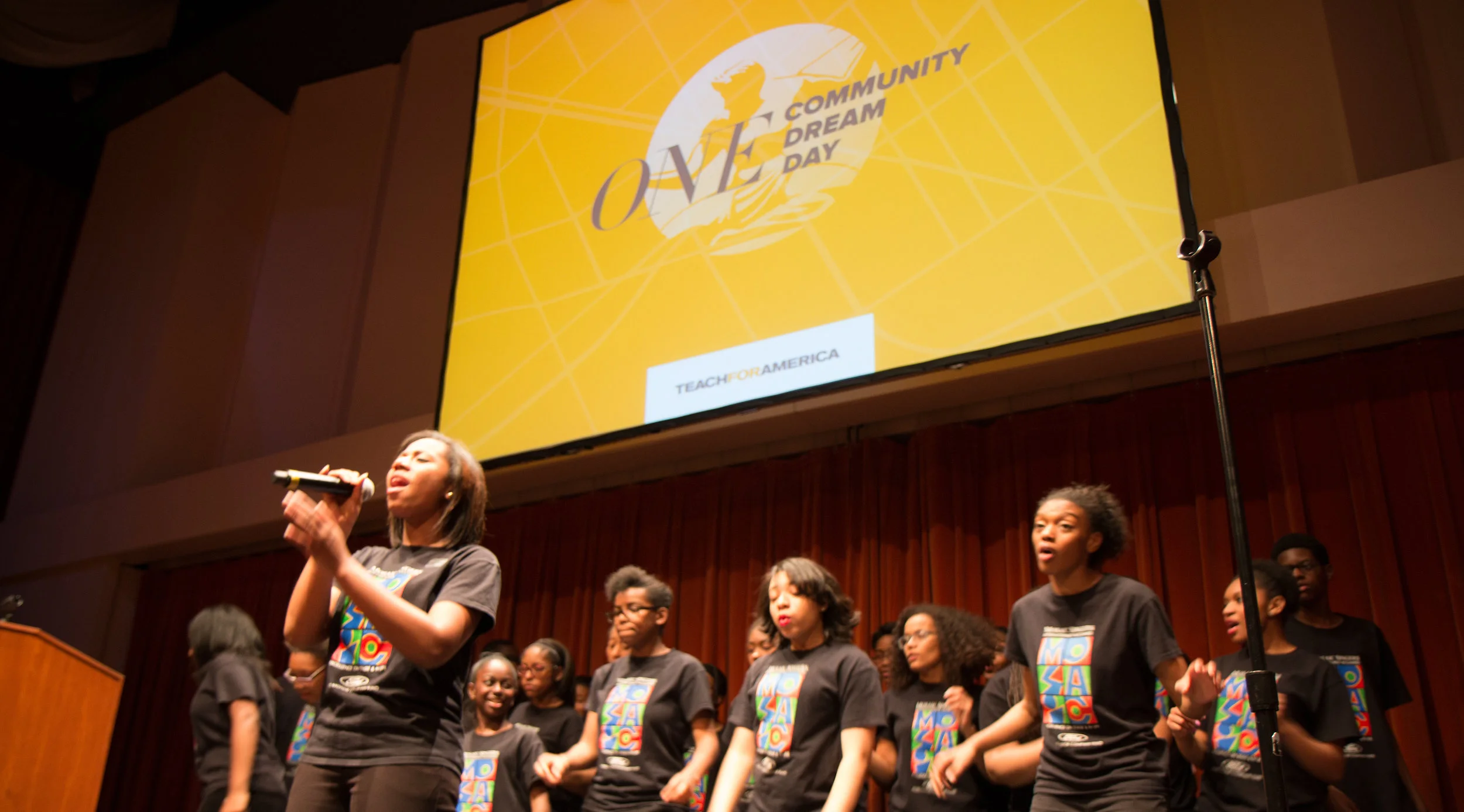 A group of young performers singing on stage, wearing matching black t-shirts with colorful designs. A large screen in the background displays "ONE Community Dream Day" and "Teach For America." The stage is set in an auditorium with a podium visible 