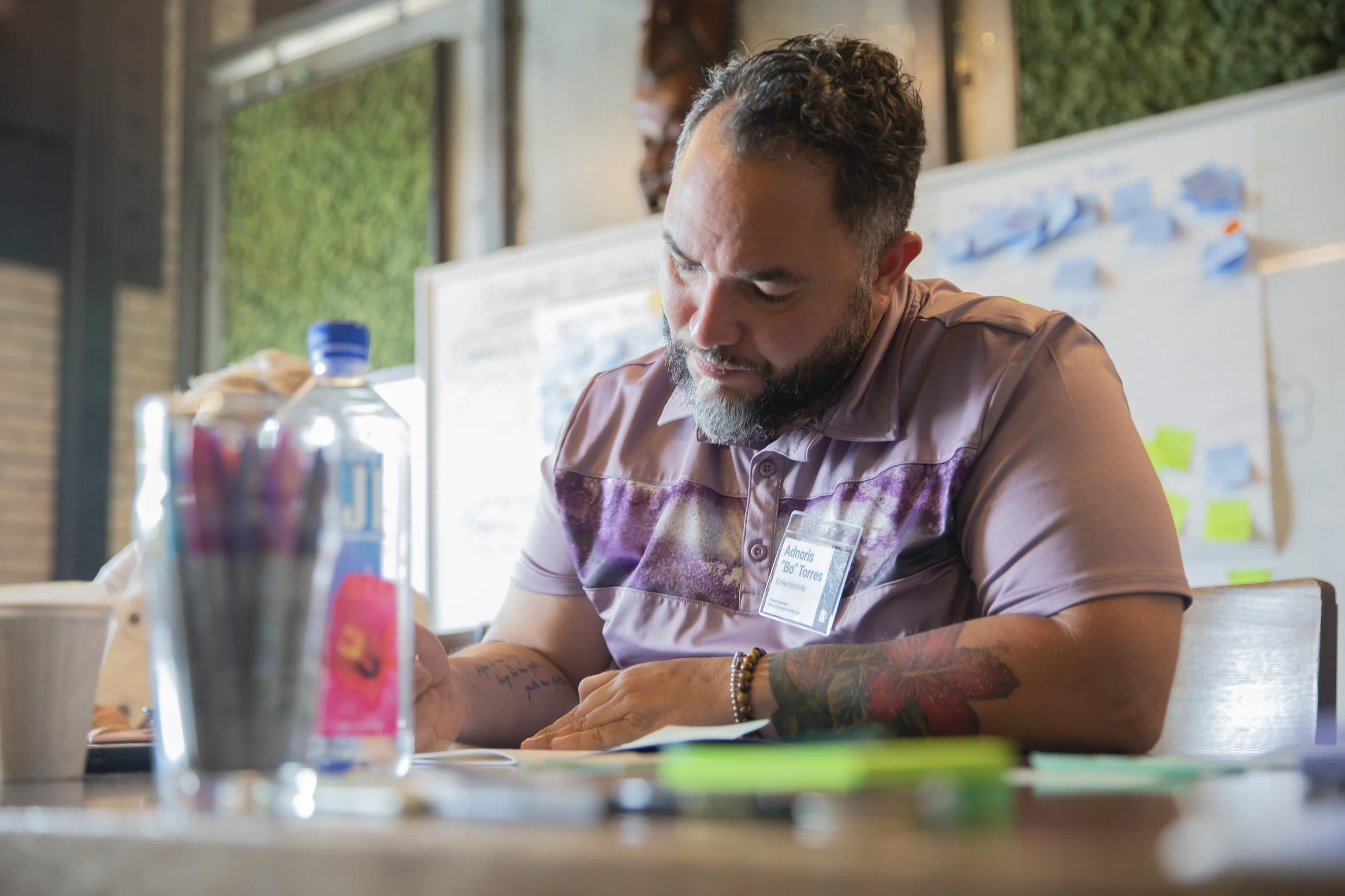 A man focusing on writing at a desk, with a water bottle and cup nearby. Office supplies such as pens and paper are on the table, and a whiteboard with sticky notes is in the background.