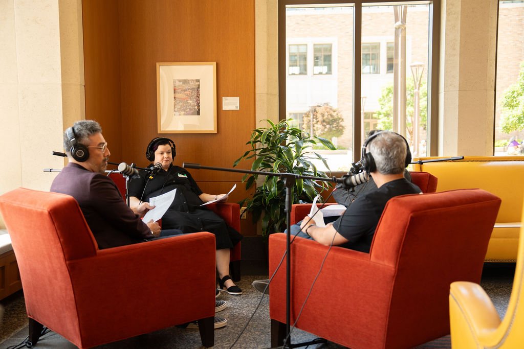 Four people seated in orange chairs during a podcast interview, wearing headphones and speaking into microphones, with a large window and potted plant in the background.