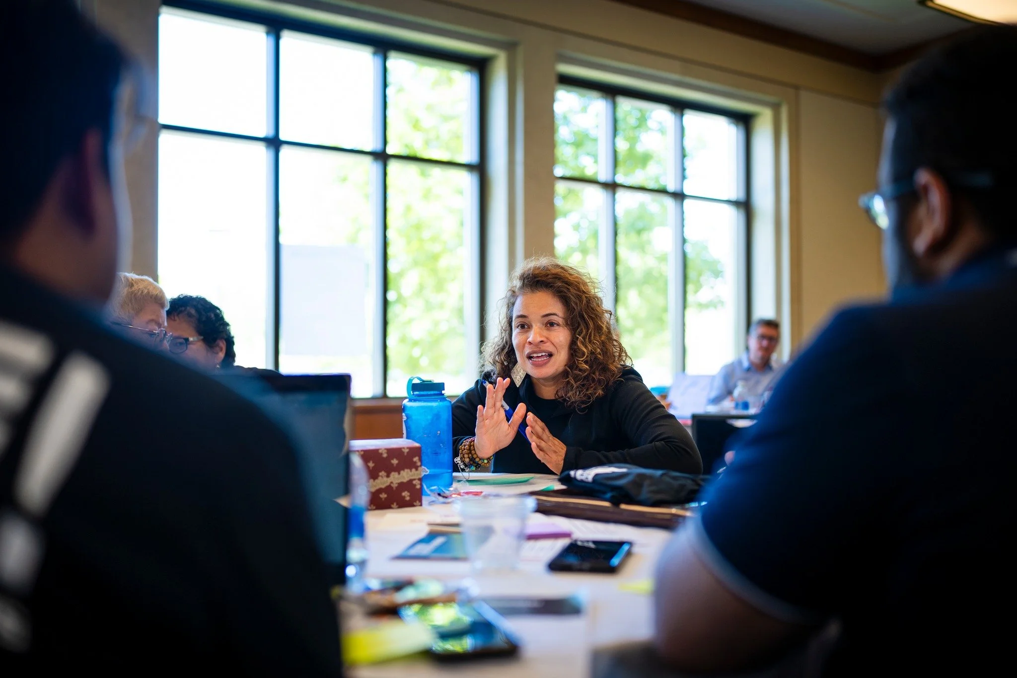 A woman with curly hair speaking to a group in a meeting room with large windows and natural light.