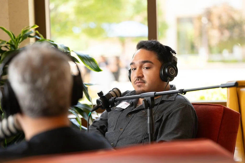 A man wearing headphones sitting in front of a microphone during a recording or interview, with a window and greenery outside.