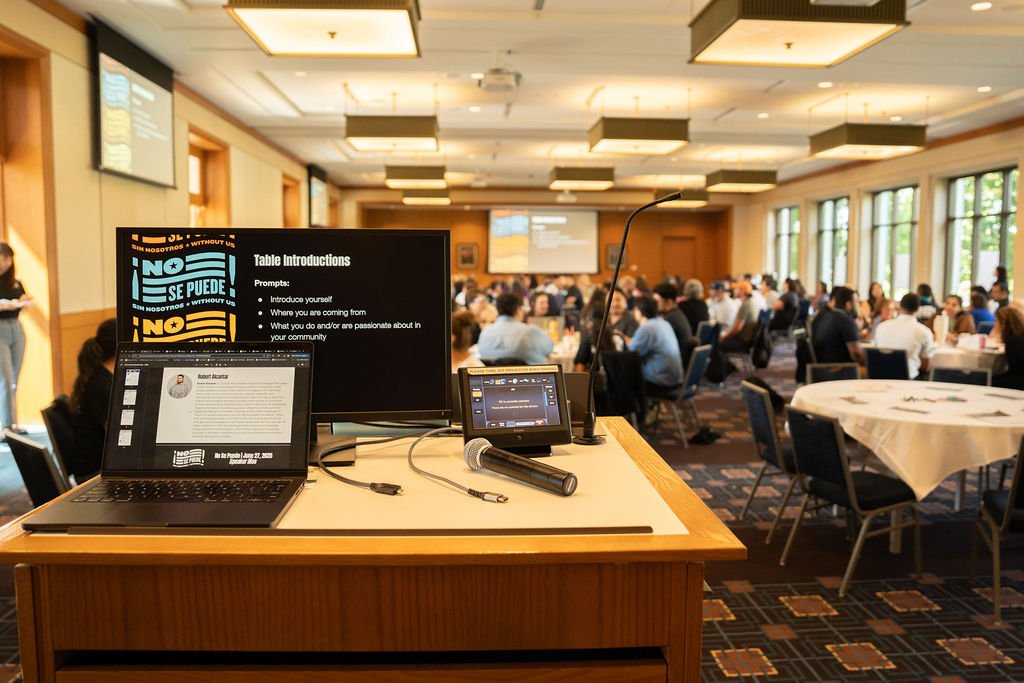 Conference room with attendees seated at round tables and a presentation setup in the foreground with a laptop, microphone, and monitors.