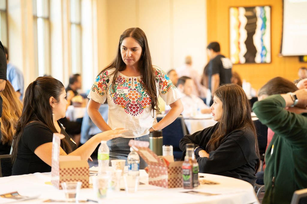 Women engaged in a discussion during a conference or workshop, with tables and materials in a well-lit indoor setting.