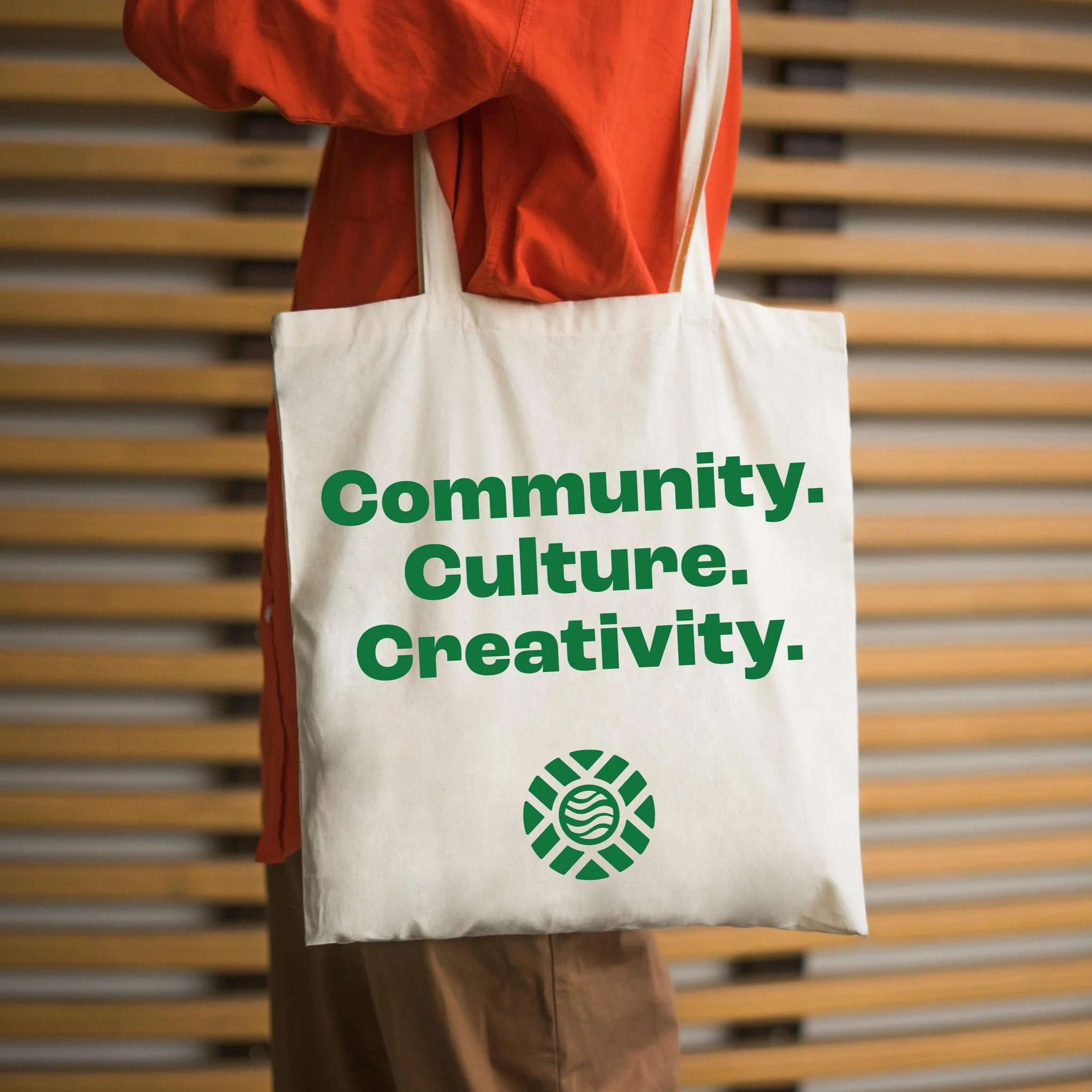 A person wearing an orange jacket holding a beige tote bag with the words 'Community. Culture. Creativity.' printed in green, and a green circular logo below.
