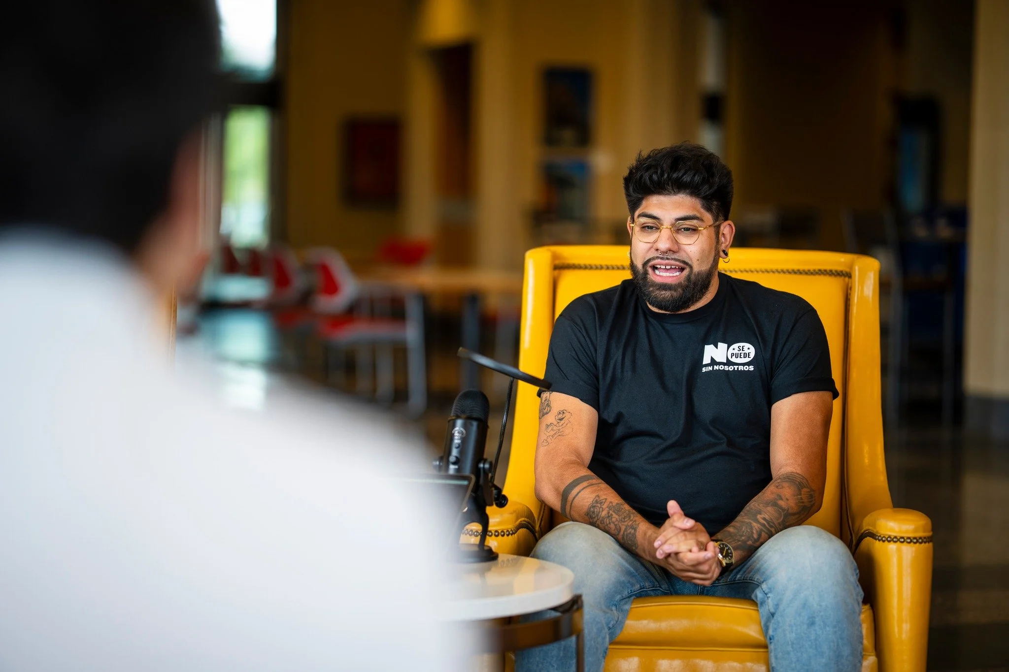 A man with glasses and tattoos talks during an interview in a room with wooden walls and red chairs.