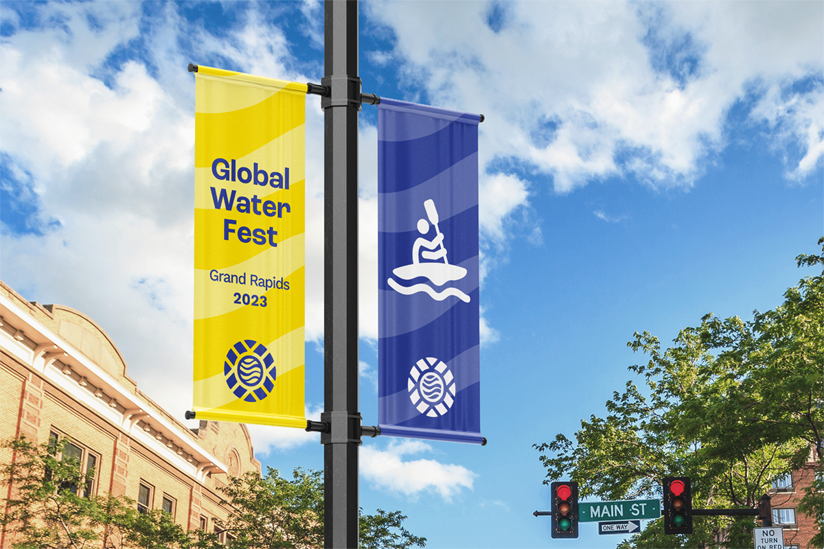 Two street banners hanging on a lamppost for the Global Water Fest 2023 in Grand Rapids, with one yellow banner and one blue banner, and a streetlight with signal lights and a sign for Main Street.