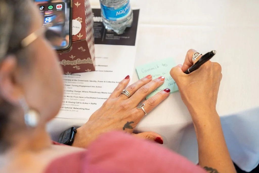 A woman with red nail polish and multiple rings writes on a sticky note with a black pen. Her hand is on a white table with a piece of paper, a water bottle, a box, and a phone visible in the background.