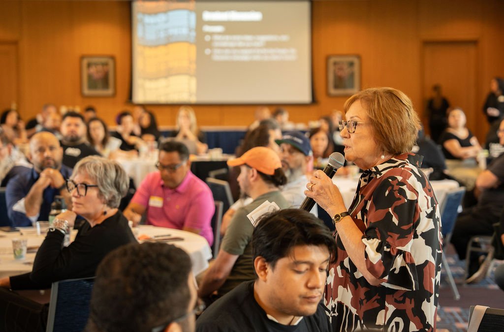 A woman with glasses and a patterned blouse is speaking into a microphone during a conference or seminar, with a large group of people sitting at round tables in a wood-paneled room.
