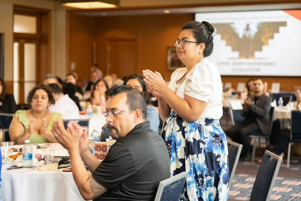 A woman in a white blouse and blue patterned skirt standing with hands clasped in applause at a formal event or conference, with attendees sitting at tables in the background.
