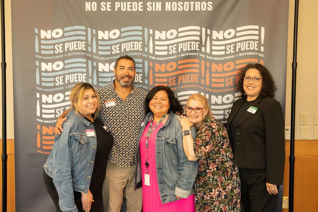 Group of five women and one man standing in front of a large banner with messages in Spanish and English that say, "No se puede sin nosotros" and "We can't without us," smiling for the photo.