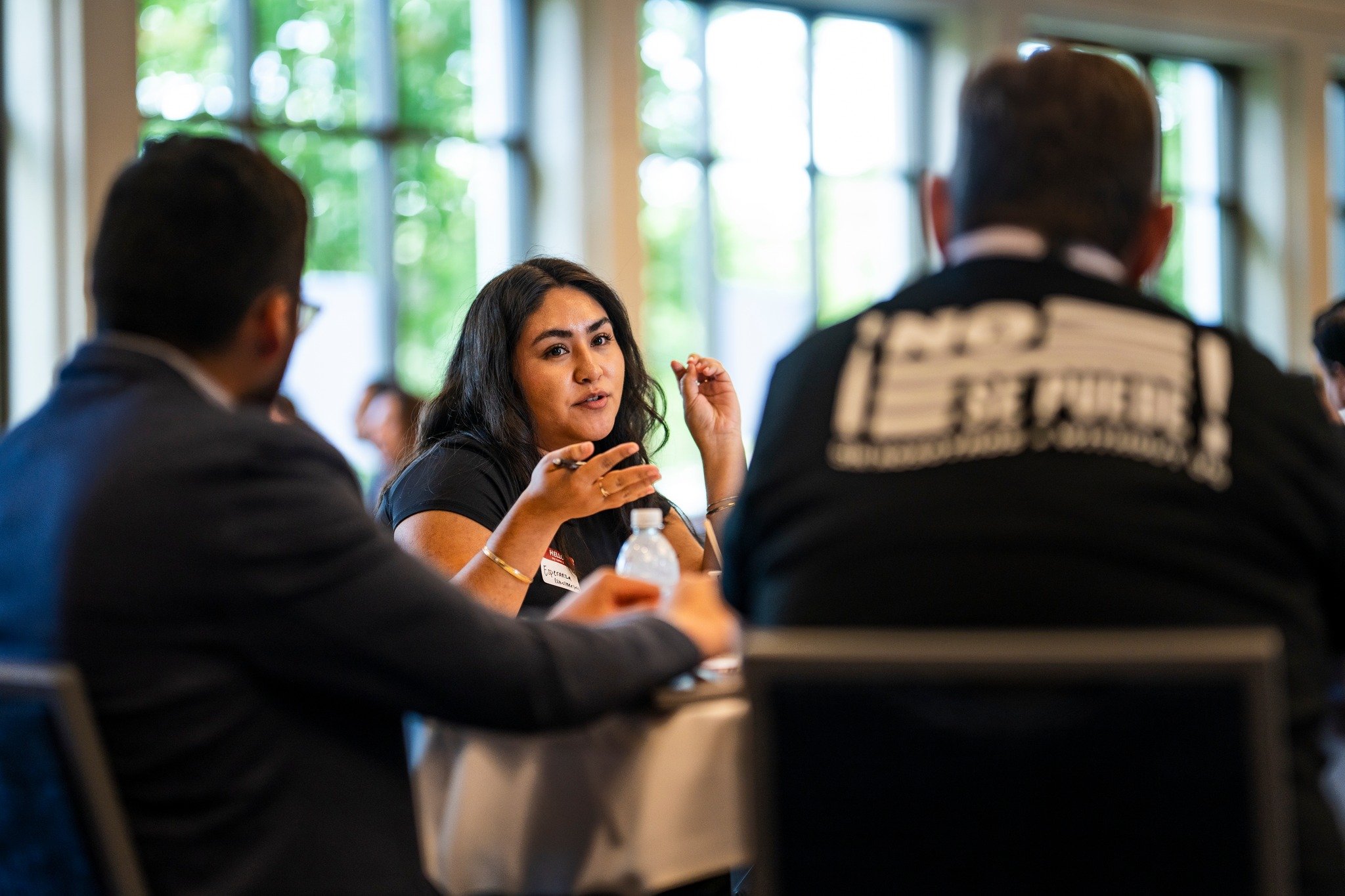 A woman with dark hair speaking passionately during a discussion at a conference or meeting, with two men in business attire listening to her.