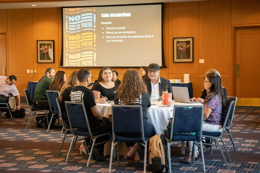 A group of diverse people seated around a round table in a conference room, participating in a discussion or workshop. A large screen displays the title "Table Introductions" and prompts for participants to introduce themselves, where they are coming