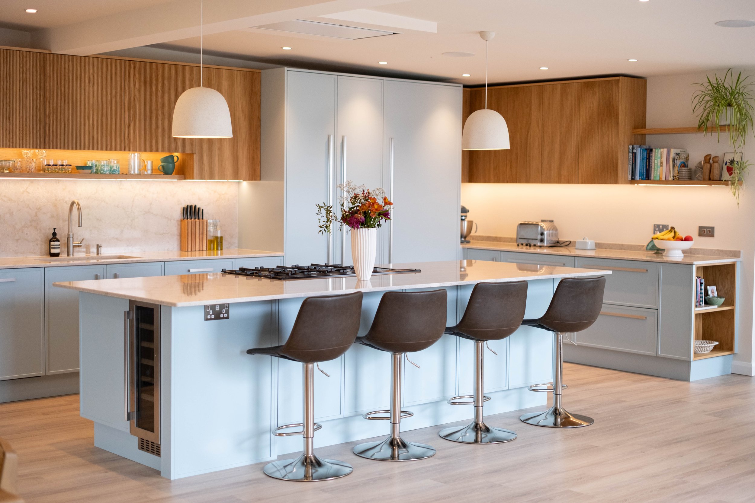 Modern kitchen with an island, light blue cabinets, wooden accents, white marble countertops, also features hanging pendant lights, bar stools, and a vase with fresh flowers.