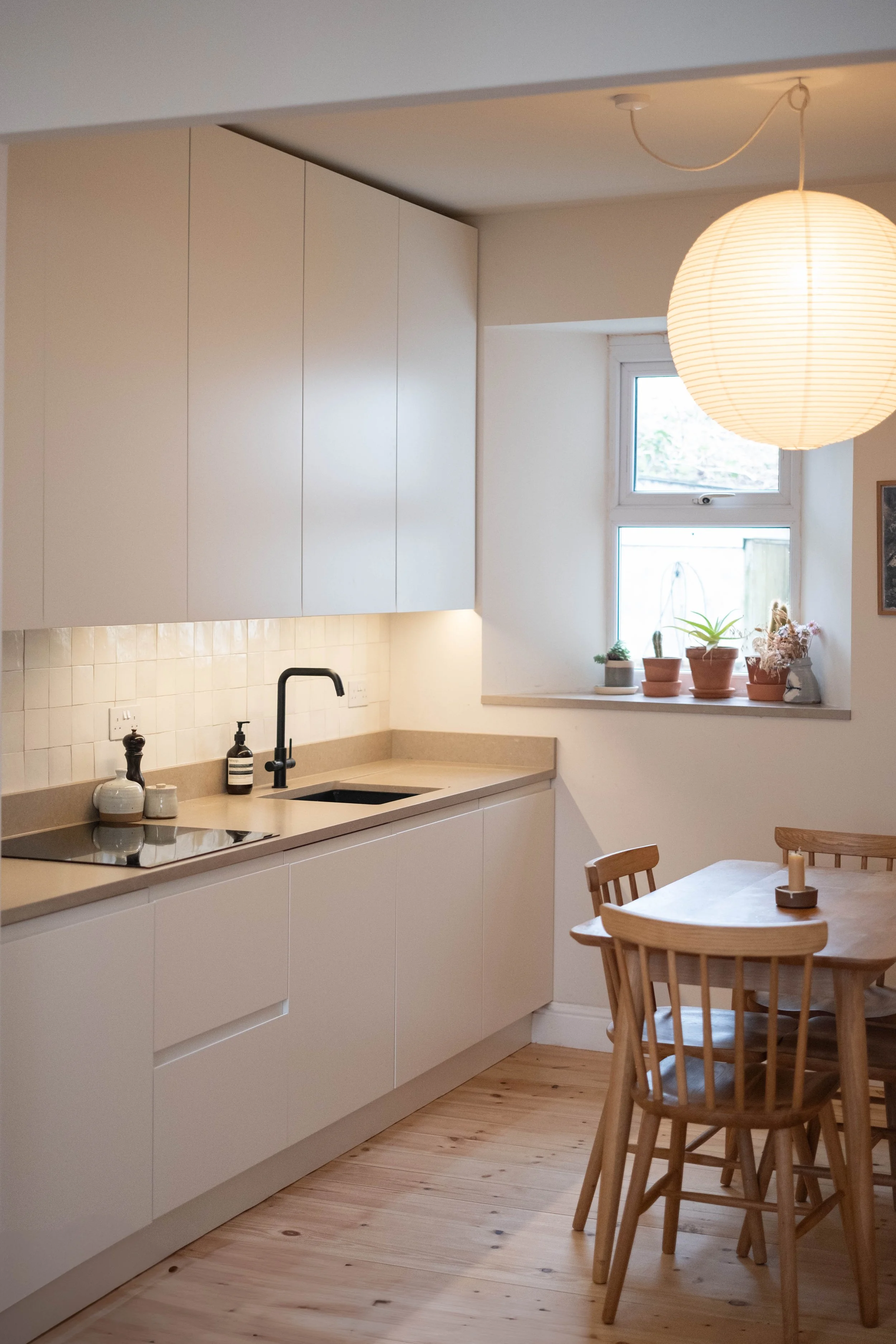 A cozy kitchen with white cabinets, wooden flooring, and a round wooden dining table with matching chairs. There is a window with potted plants on the sill, and a paper lantern hanging from the ceiling.