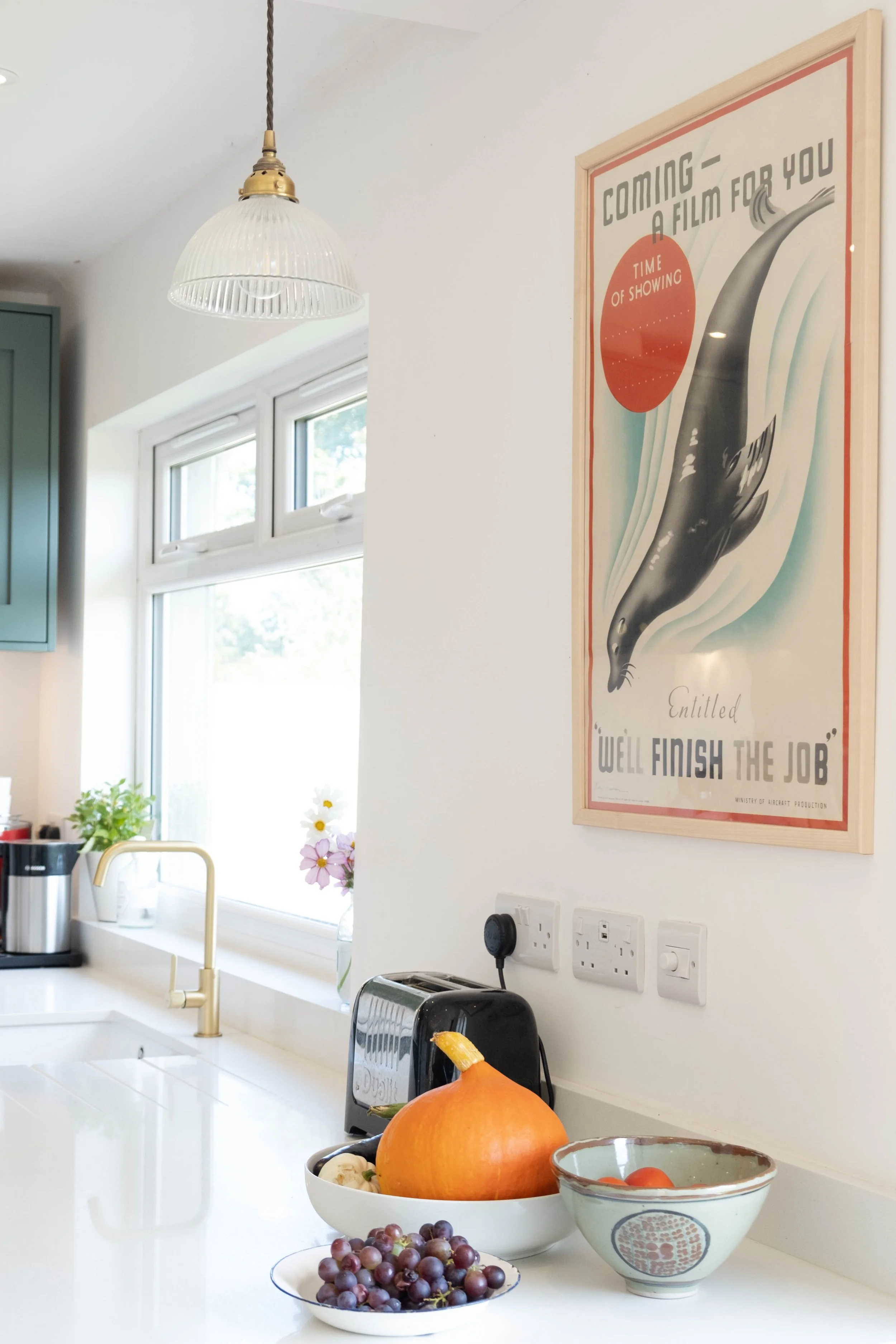 Kitchen with white counter, gold faucet, window, potted plant, pumpkin, bowls of grapes, tomatoes, and potatoes, and a framed poster on the wall that says 'Coming - A Film For You, Well Finish The Job'.
