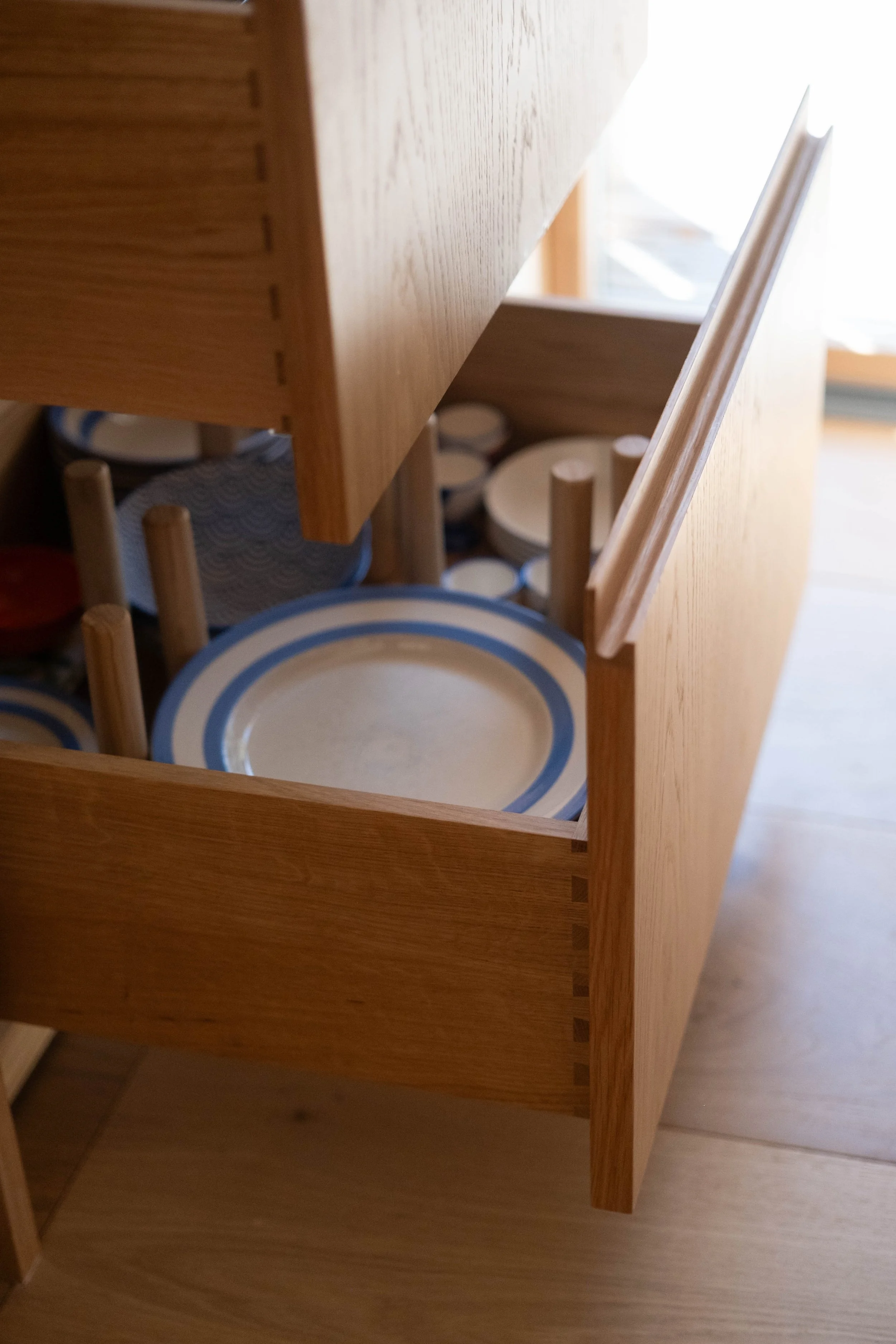 A wooden cabinet tilted sideways, with a view of plates stored inside at the bottom, and a window in the background.