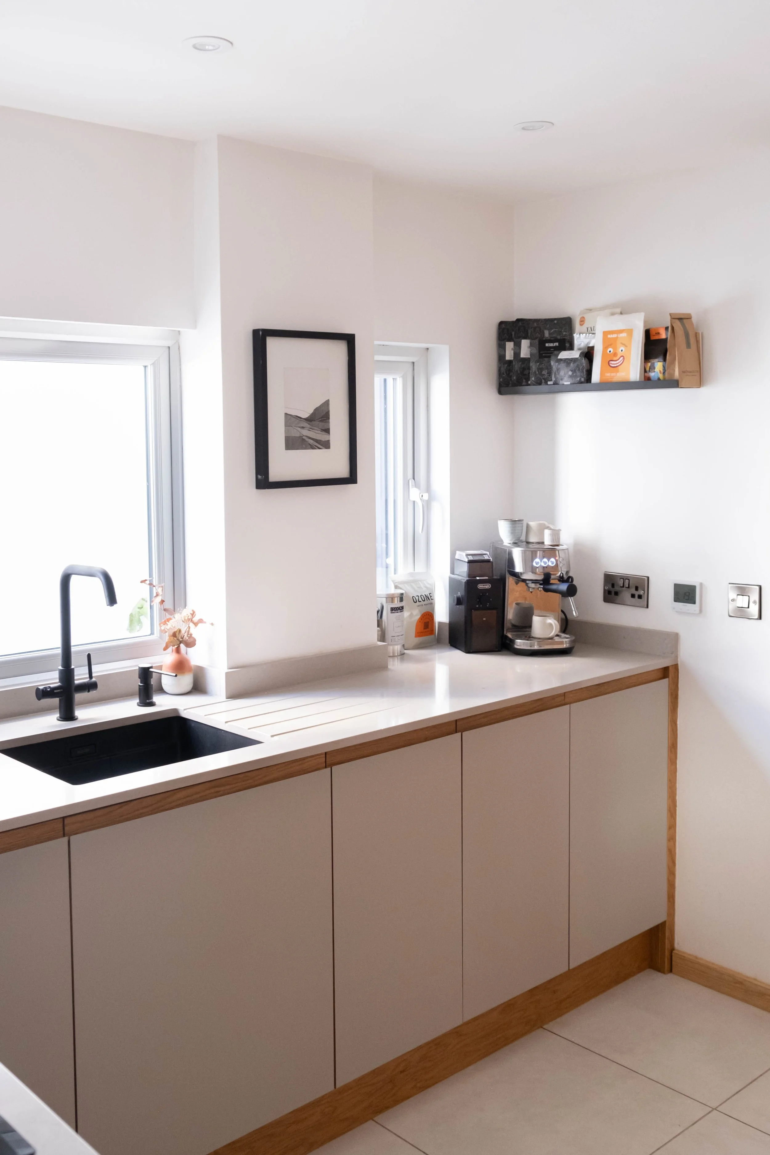Modern kitchen with white cabinets, black sink, and a window. A small potted plant is near the sink, and appliances such as a coffee machine are on the countertop. A framed black-and-white artwork hangs on the wall, and a floating shelf holds various