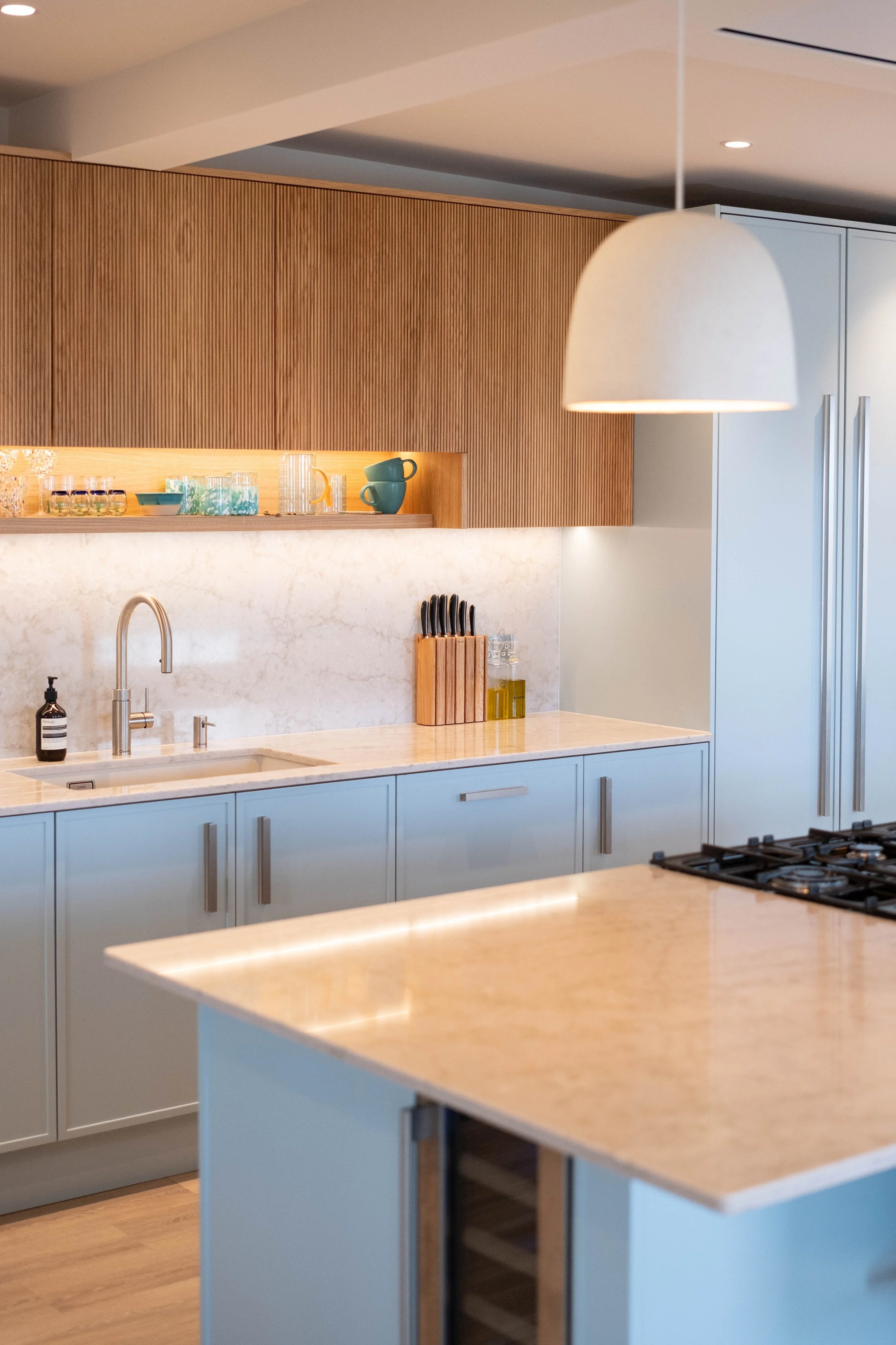 A modern kitchen with light blue cabinets, a marble countertop, and a built-in stove. There are wooden cabinets above, a hanging pendant light, and a small open shelf with glassware and cups. Additionally, there is a knife rack and bottles of oil on 