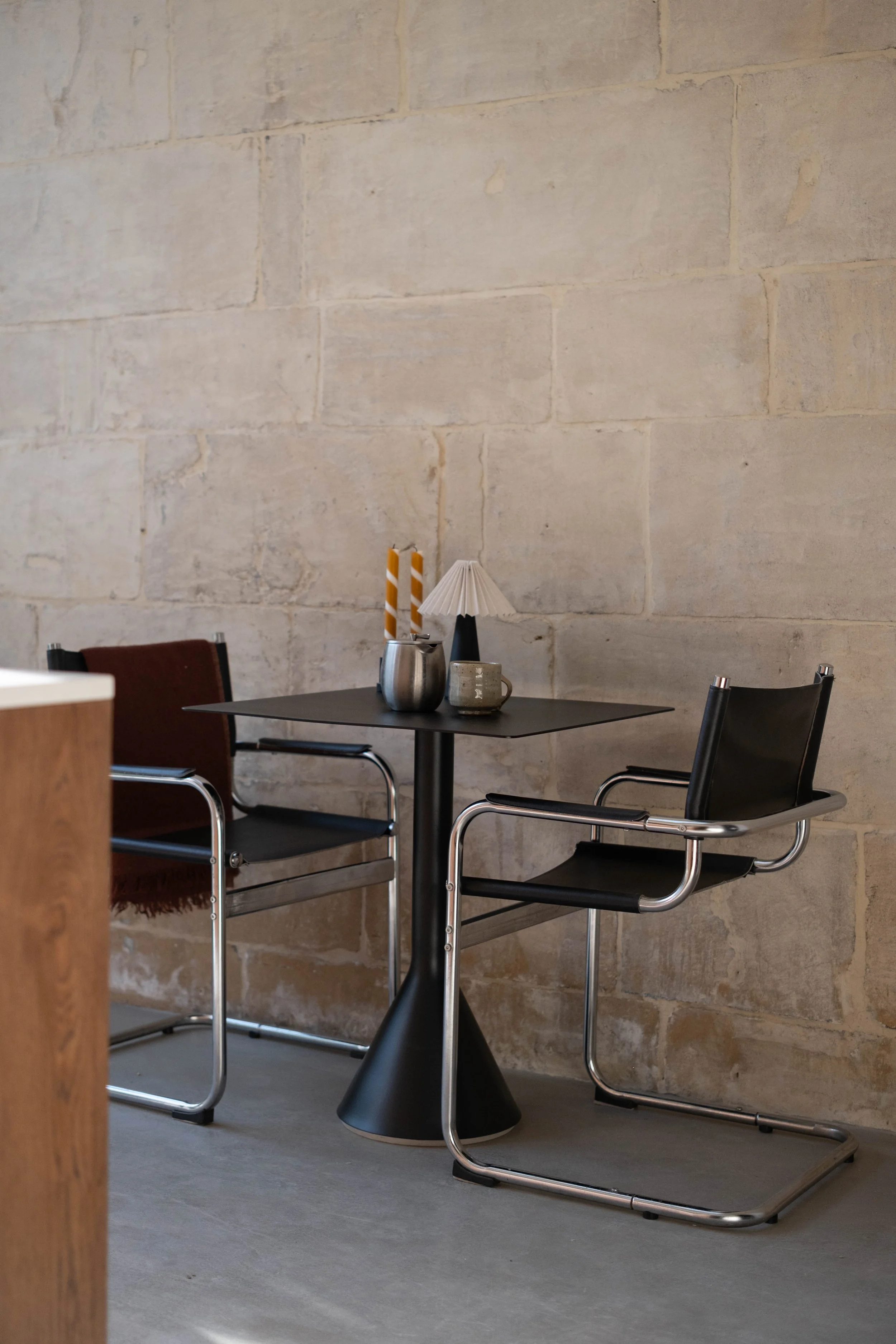 A table with a lamp, two yellow striped candles, and a mug, set against a beige stone wall with chairs around it.