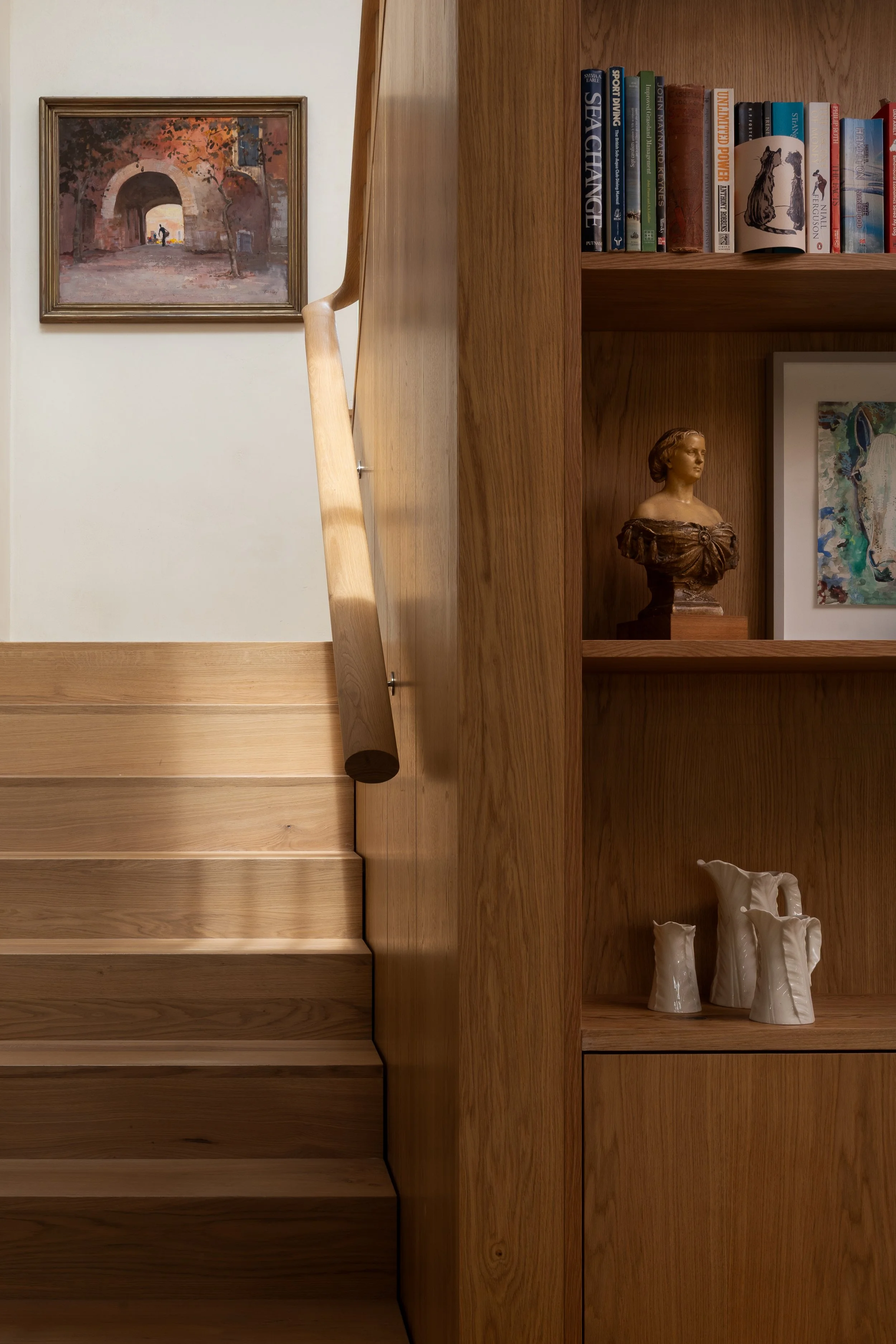 A wooden bookshelf with several books on top and a small sculpture of a woman's bust inside the compartment. A framed painting of an archway, trees, and a person is on the wall behind the bookshelf. There are also vases with abstract designs on the l
