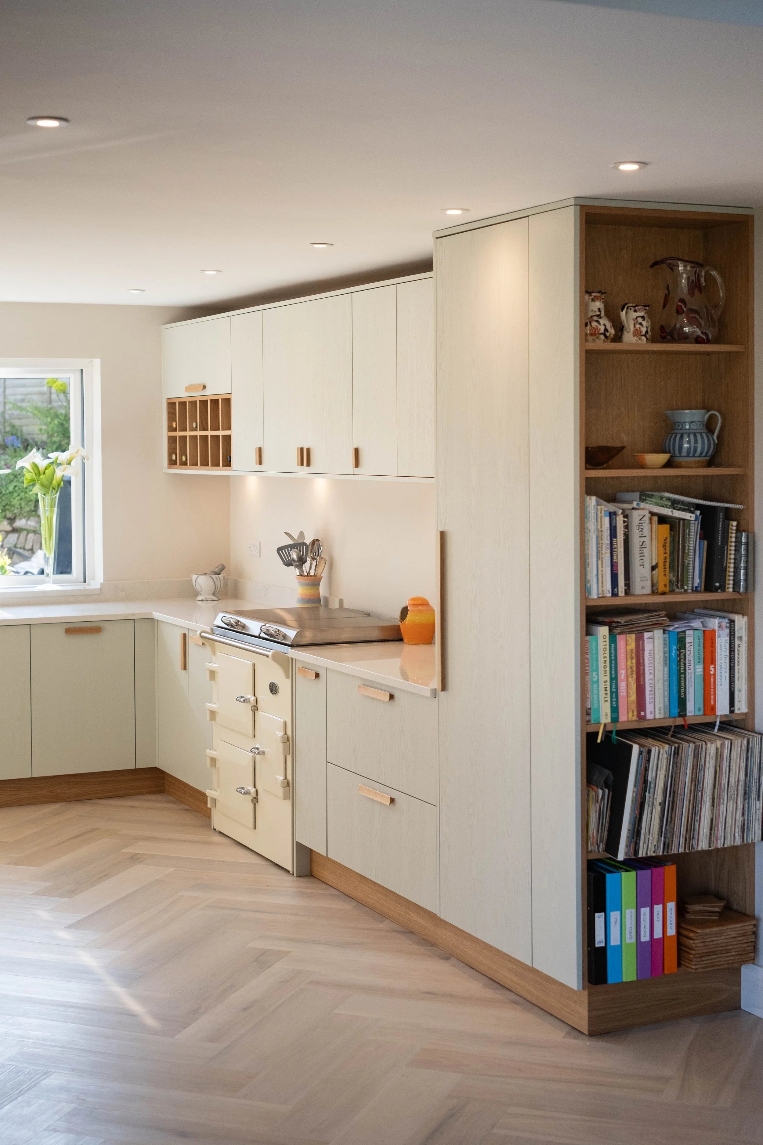 A corner of a modern kitchen with white cabinets, a window with a vase of white flowers, a stove, and a wooden bookshelf filled with books and decorative items. The floor is light wood.