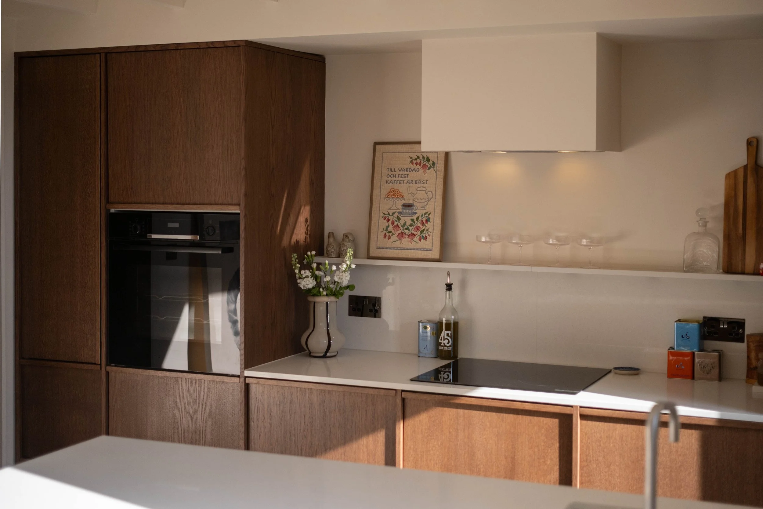 A modern kitchen with wooden cabinets, white countertops, and electrical outlets. Decor includes framed textile art, a white vase with flowers, two canisters, and a glass jar. Five wine glasses are on a shelf above the counter.