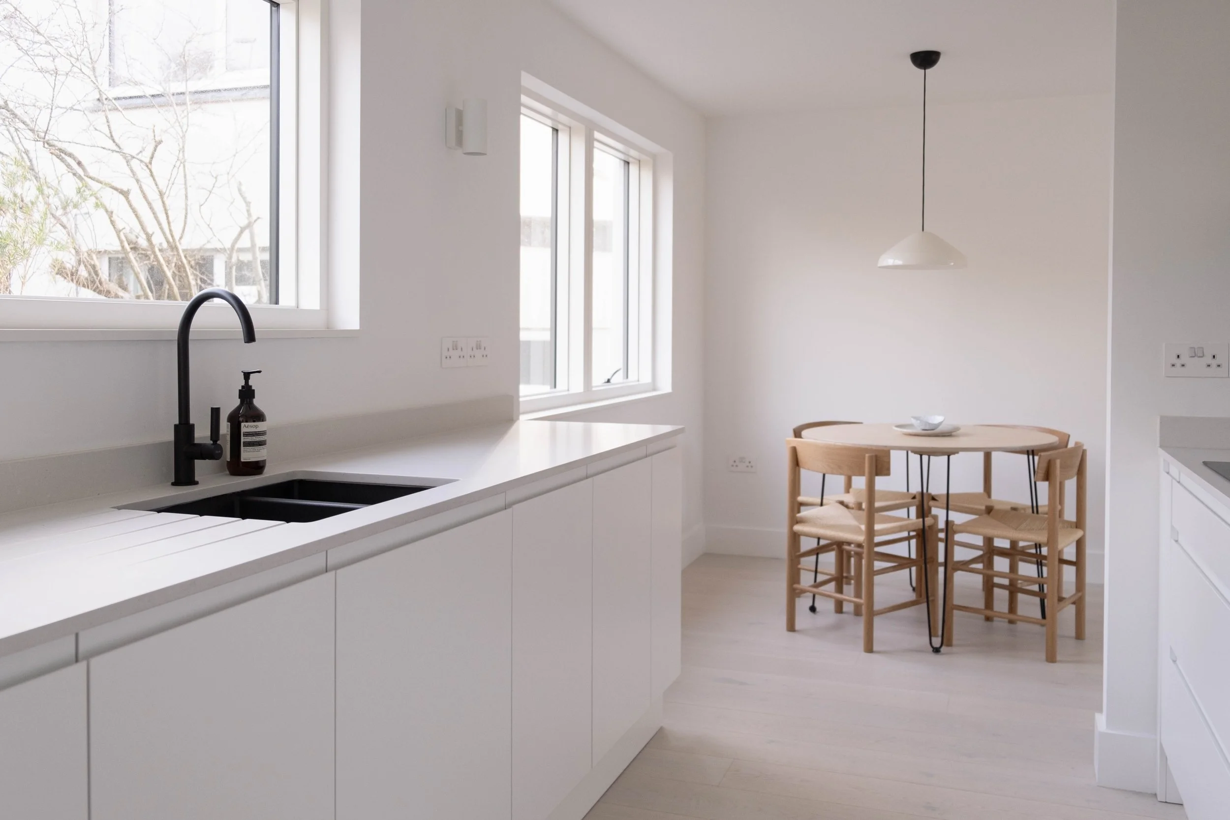 A minimalist kitchen with white cabinets, a black faucet, a soap dispenser, and a white countertop near a window. Adjacent to the kitchen is a small round wooden table with four matching chairs and a white bowl on top. There are two electrical outlet