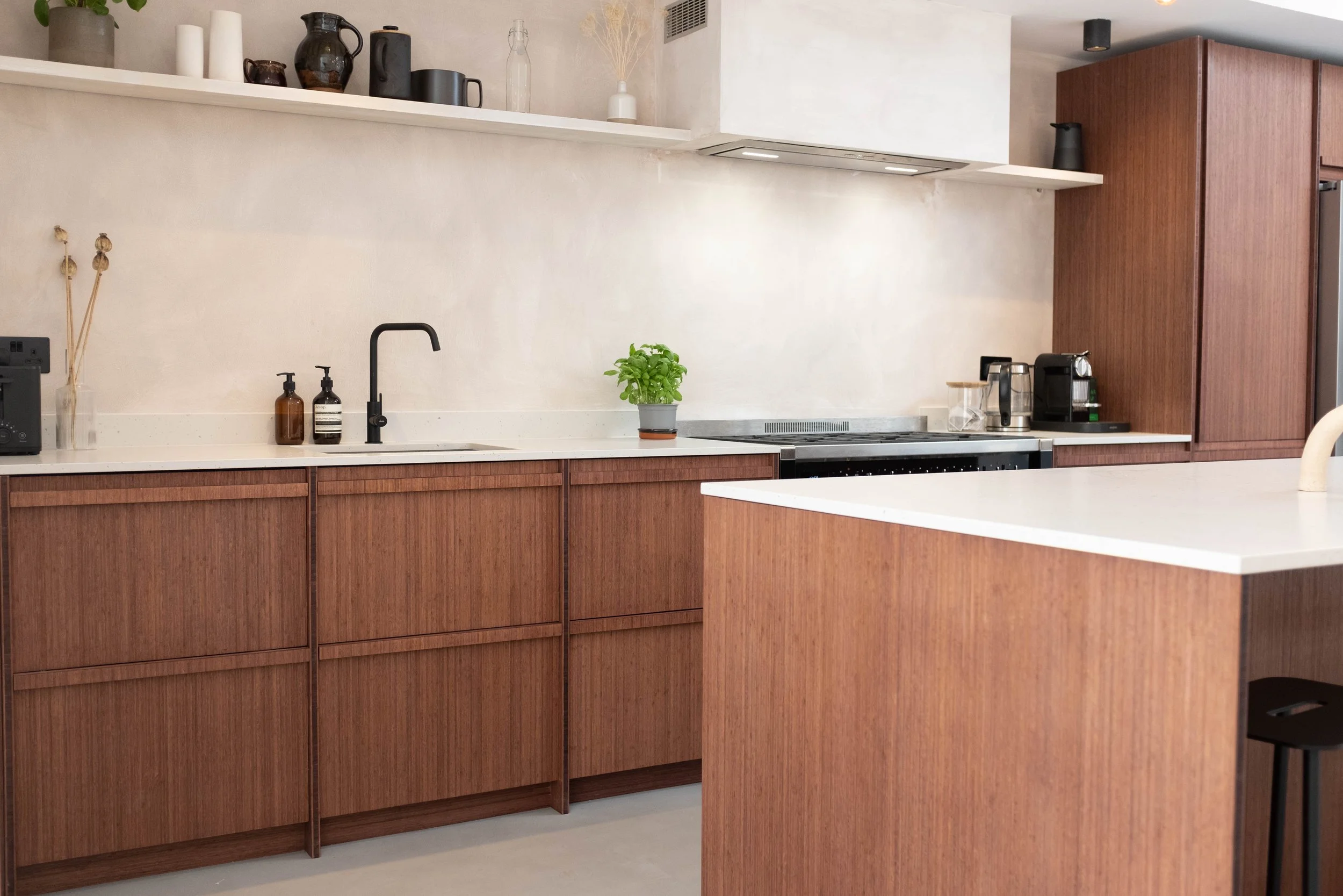 A modern kitchen with wooden cabinets, a white countertop, and potted plant. Shelves with decorative vases and kitchen appliances are visible.