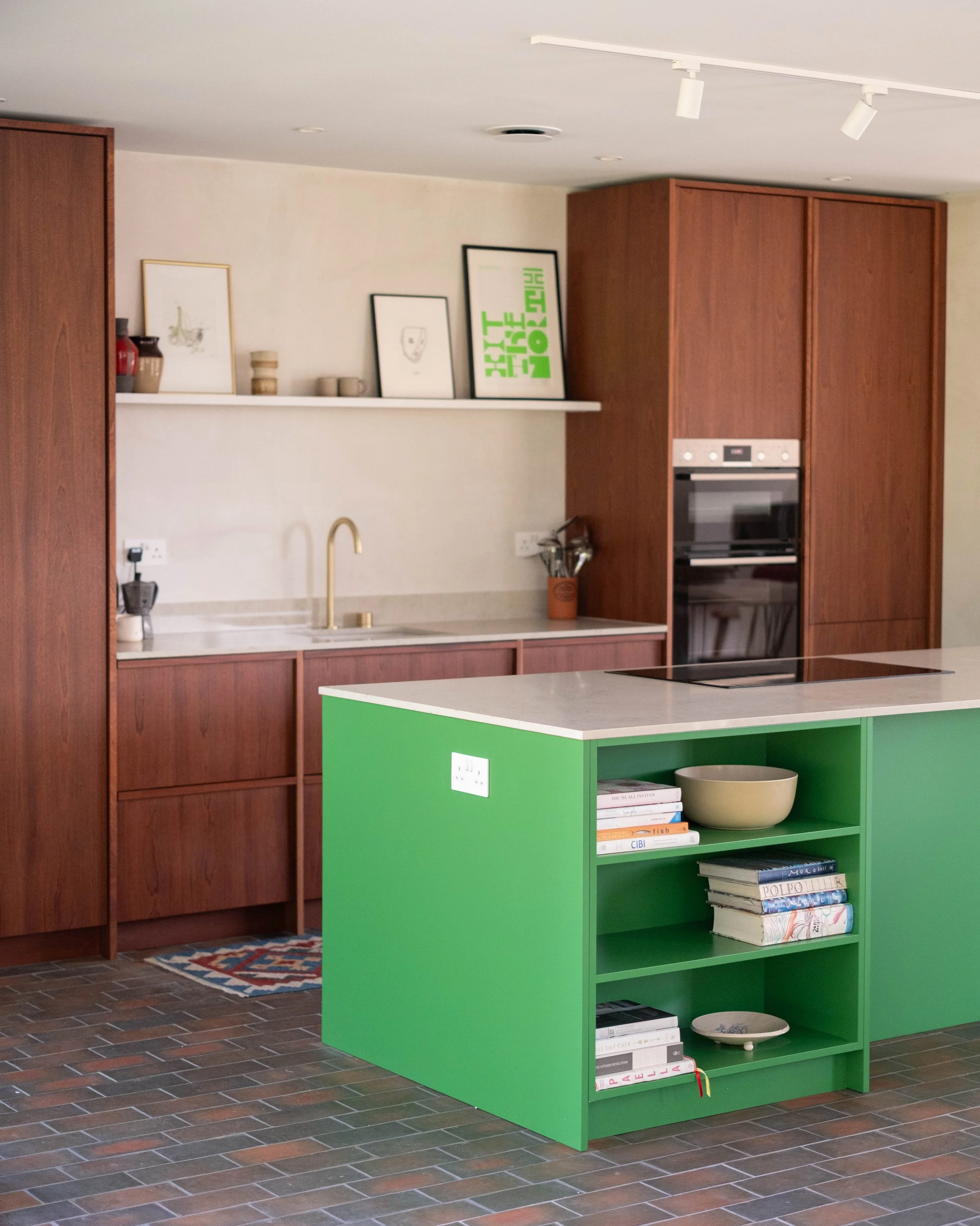 Open-plan kitchen with green island, wood cabinetry, brick floor, wall shelf with framed art, books, and decorative items.