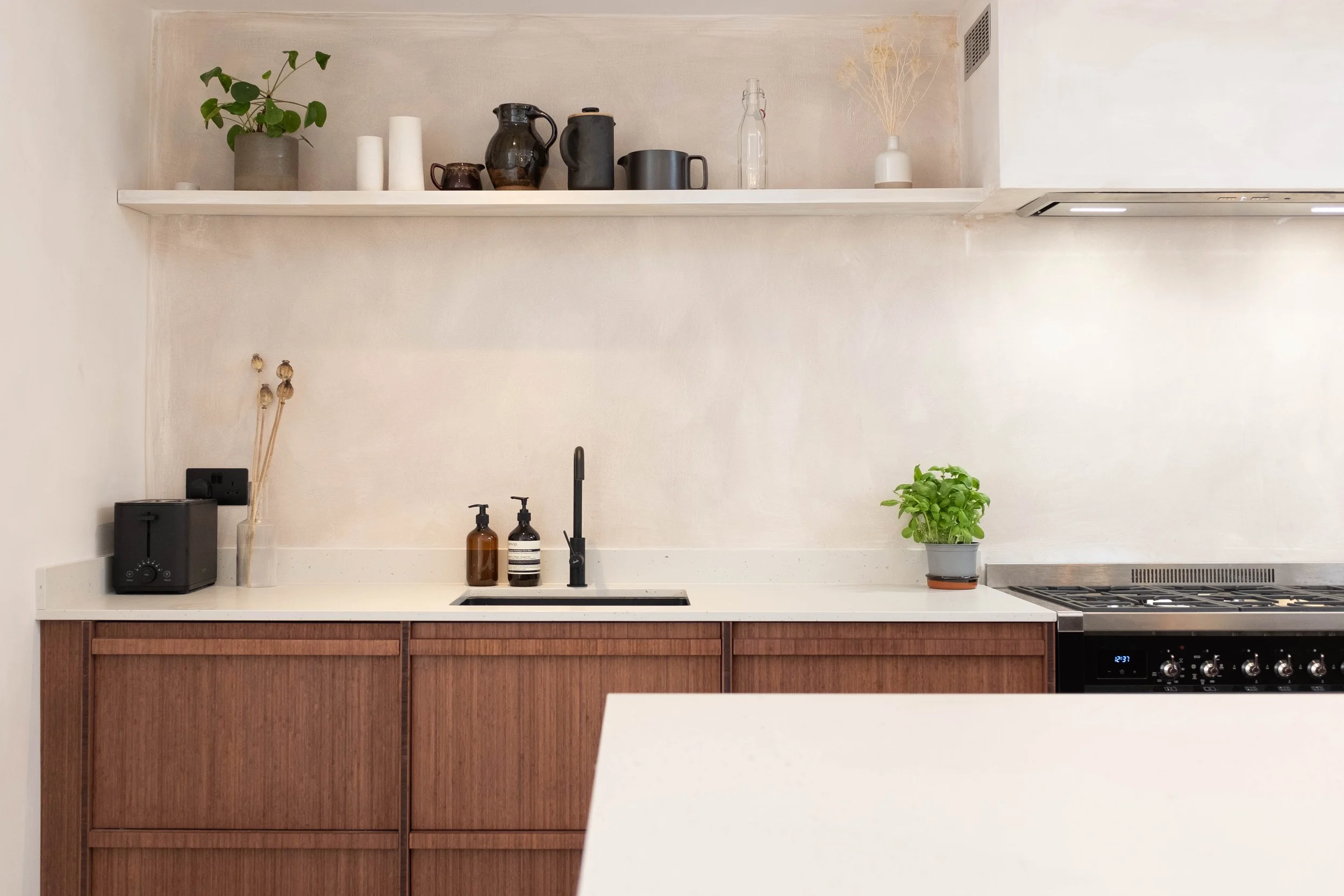 Minimalist kitchen with a white countertop, brown wooden cabinets, a black faucet, two brown soap dispensers, a small black appliance, decorative vases, and a potted plant on the counter. There is a shelf with various vases and a small plant, and par