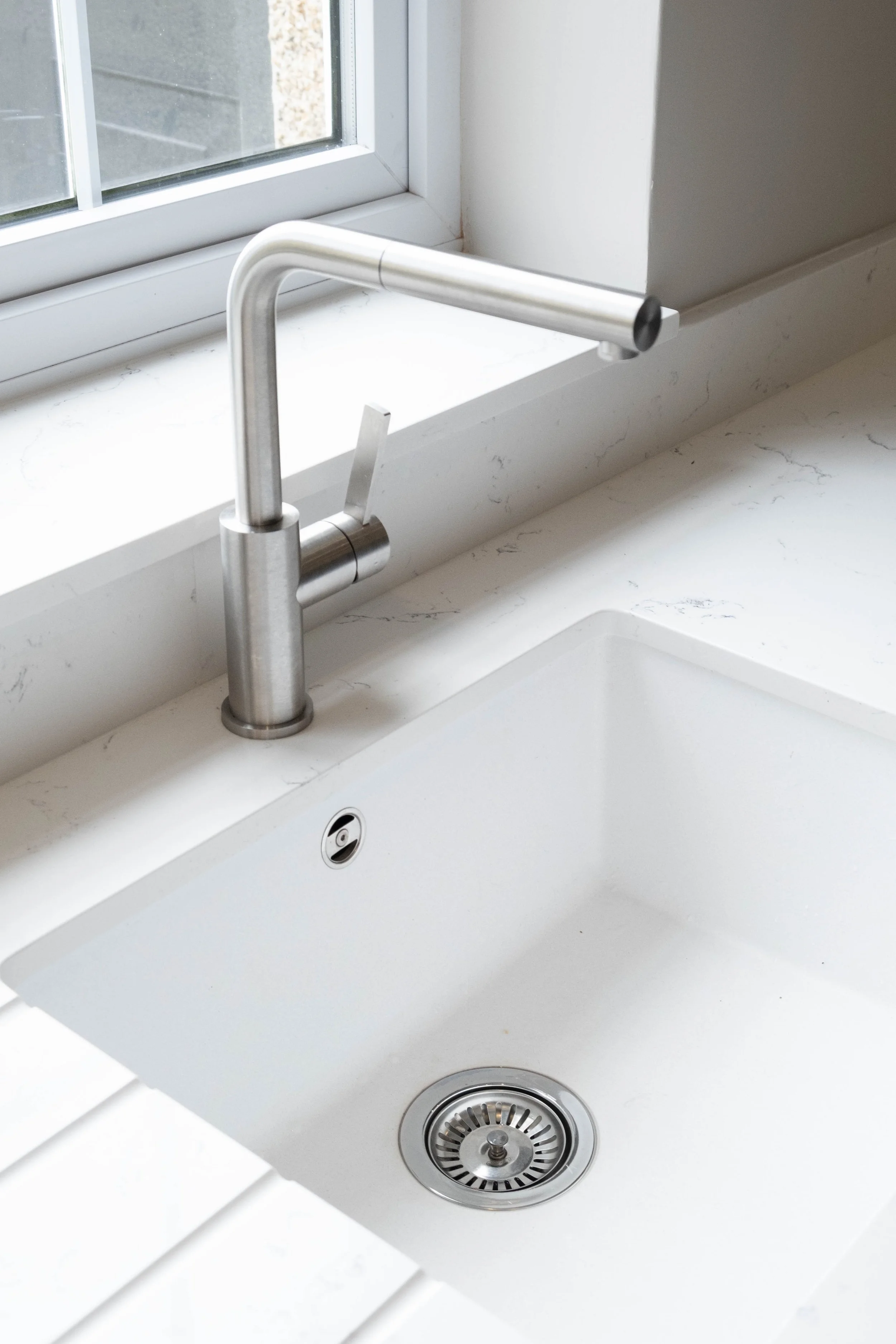 Modern kitchen sink with stainless steel faucet, white marble countertop, and window with a view outside.
