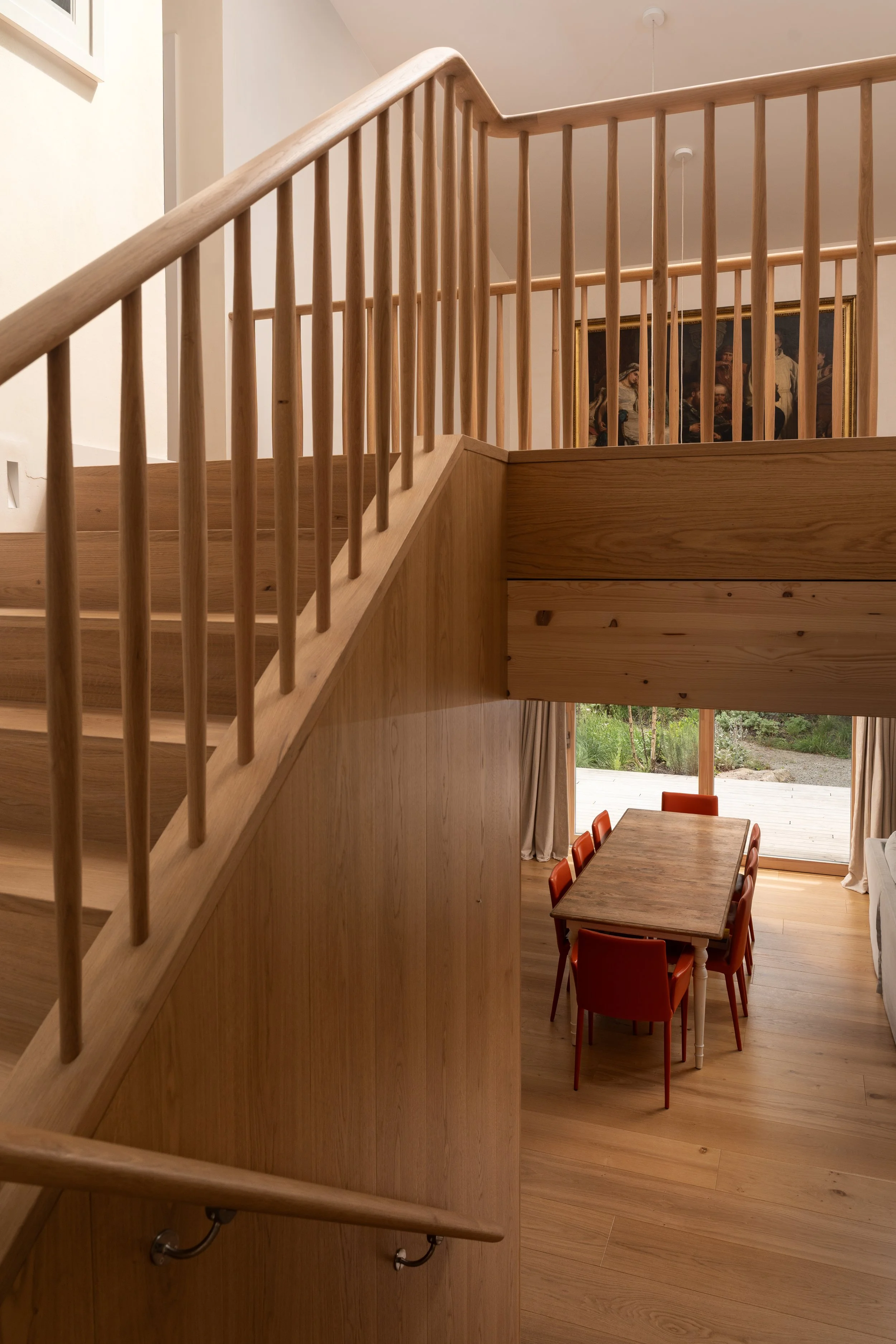 Interior view of a modern wooden staircase leading to a dining area with a table and red chairs. Large window/open door showing greenery outside, framed painting on the wall.