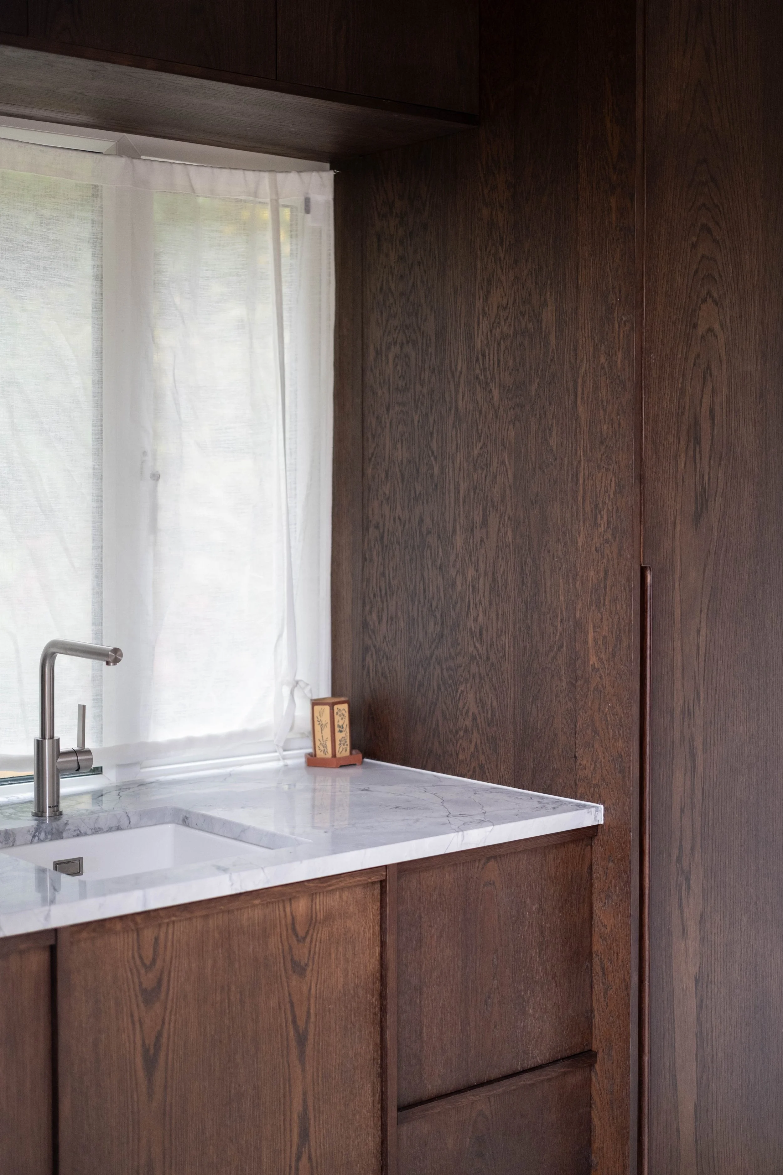 A kitchen corner with a white marble countertop, a silver faucet, dark wooden cabinets, a window with white curtains, and a small decorative object on the counter.