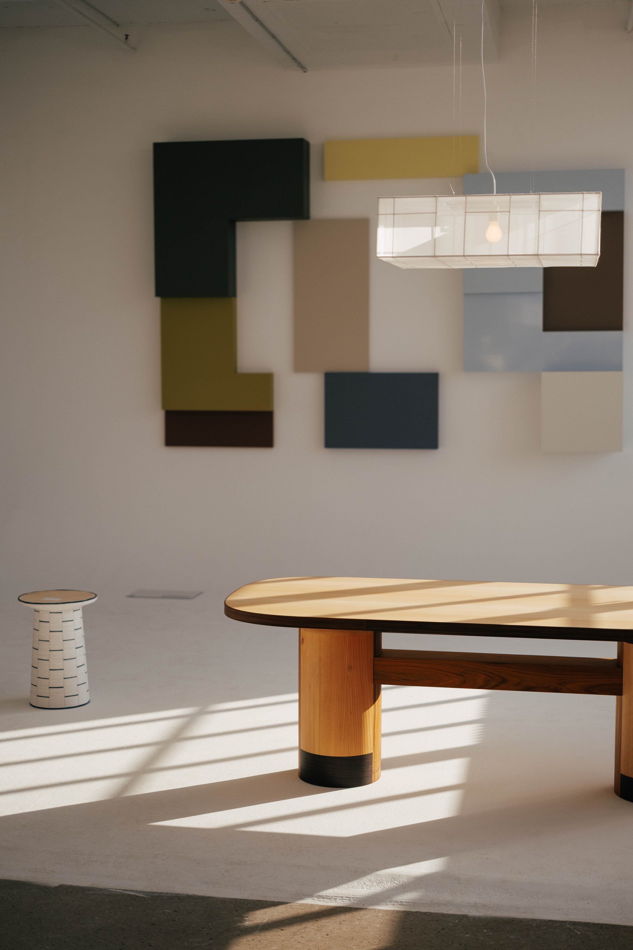 A modern interior space with a wooden table, a white and black striped stool, and a wall decorated with colorful square panels and a suspended light fixture. Sunlight casts shadows on the floor.
