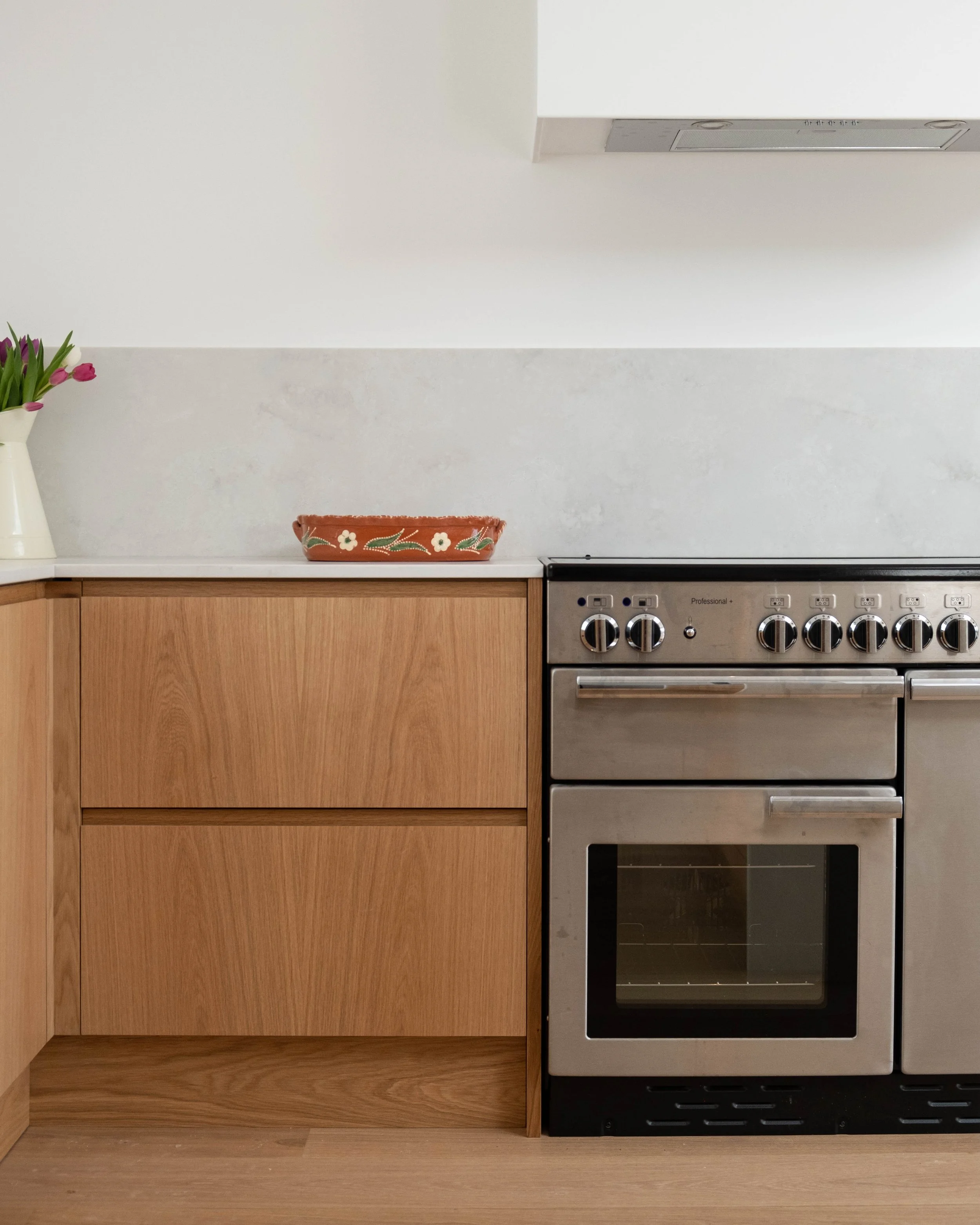 A kitchen countertop with a wooden cabinet below and a stainless steel oven with black glass doors. On the countertop, there is a white vase with pink tulips on the left and a decorative terracotta bowl with white flowers and green leaves in the midd