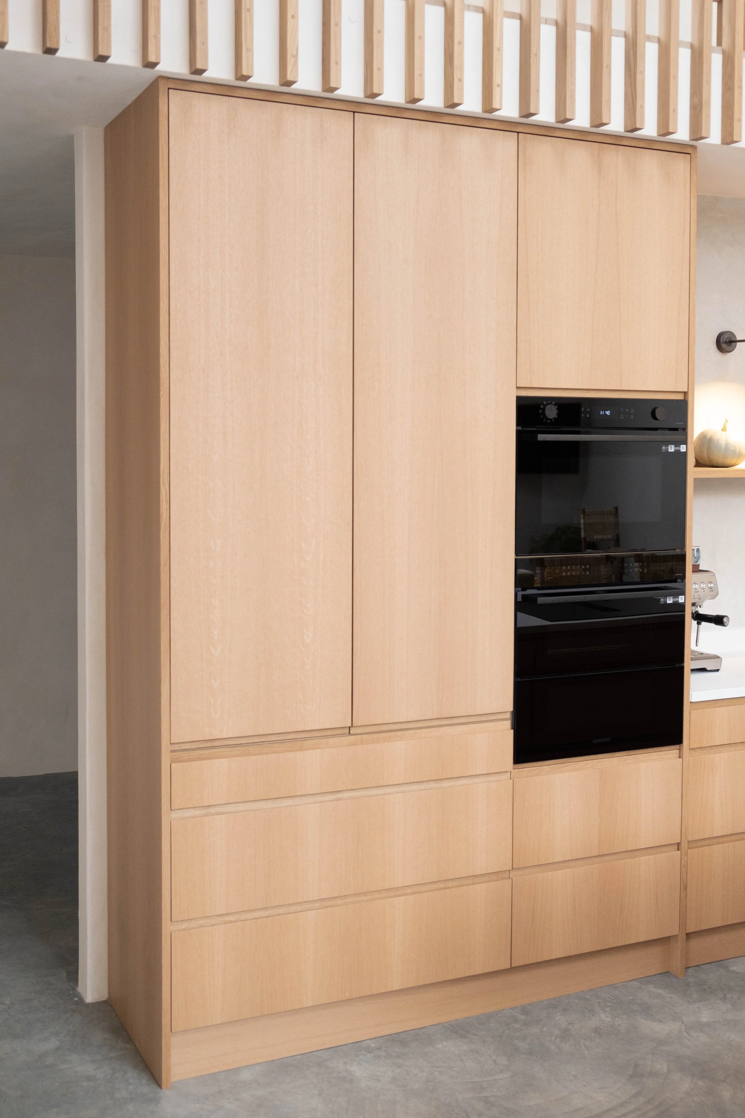 A large built-in wooden cabinet with closed doors and drawers, containing a black oven and microwave, in a modern kitchen with light-colored walls and a grey floor.