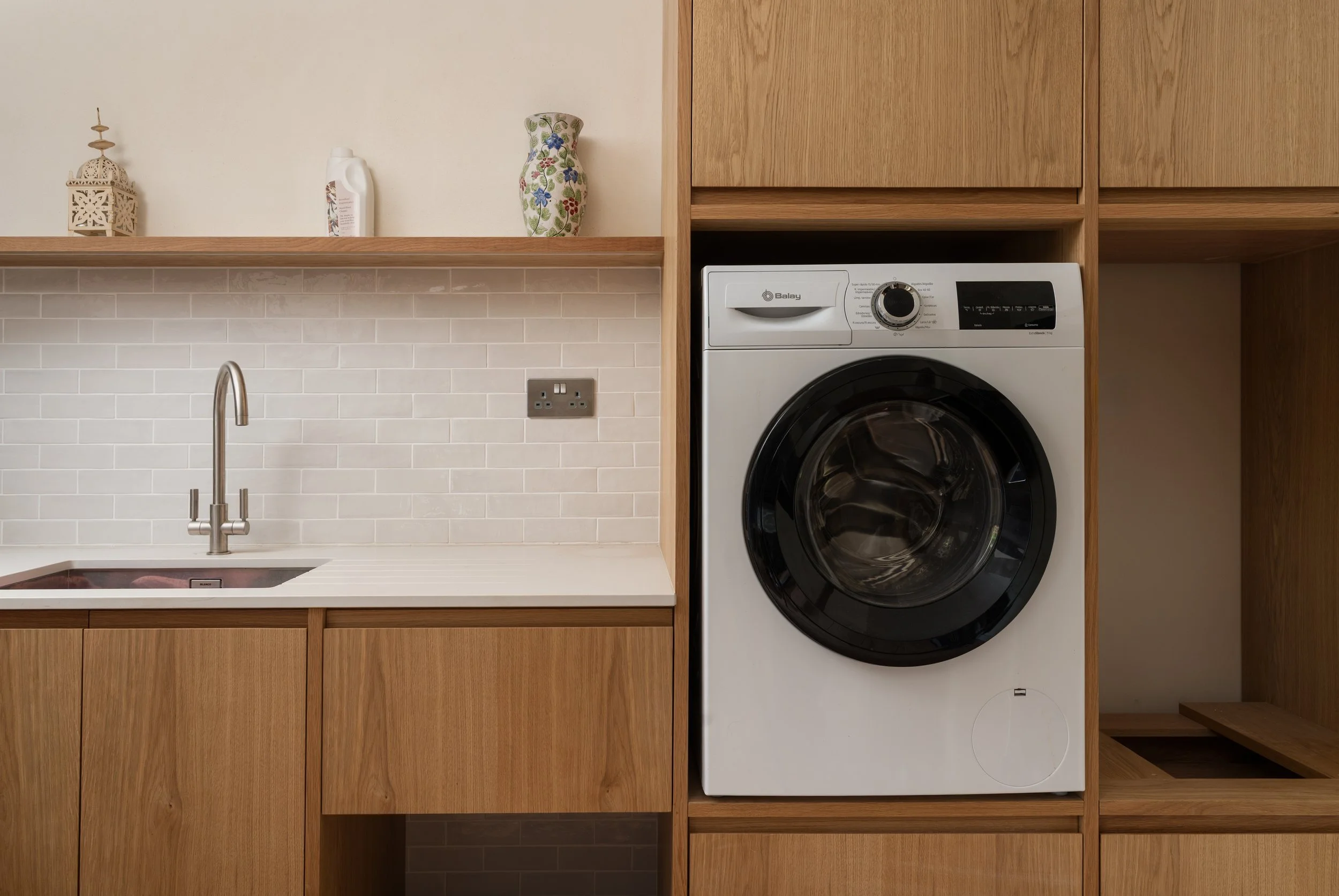 A laundry room with wooden cabinets, a white washing machine, a stainless steel sink with a faucet, a decorative lantern, a floral vase, and a bottle of detergent.