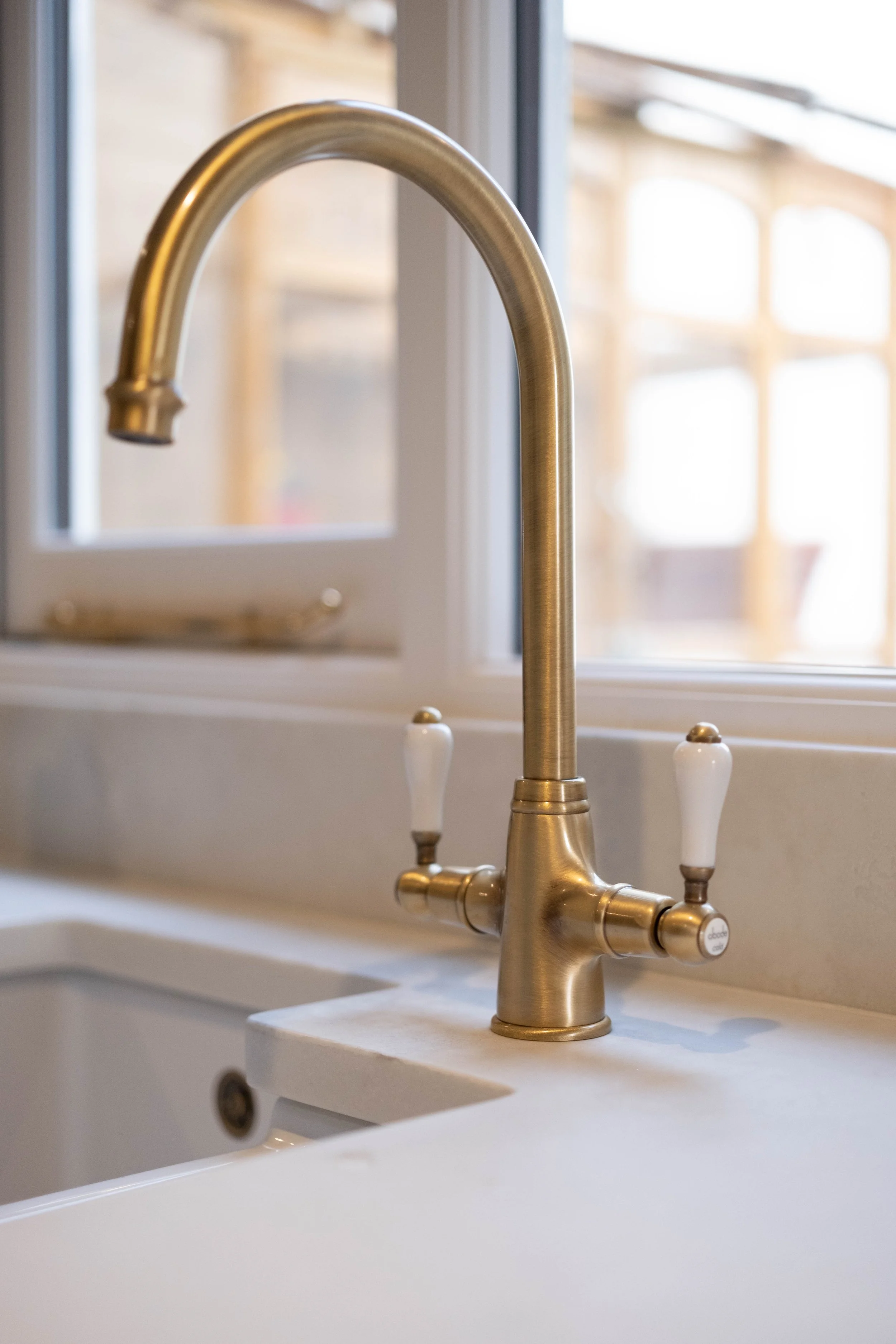 A kitchen sink with a brushed brass faucet and white handles, set in a white countertop near a window with bright sunlight streaming in.