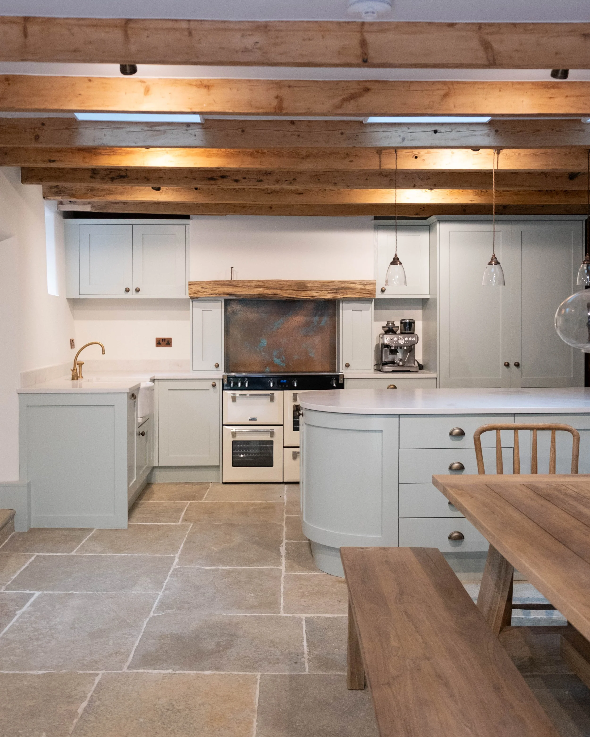 Interior of a kitchen with wooden ceiling beams, white cabinets, a stove, a countertop, and a wooden table and benches. Pendant lights hang from the ceiling.