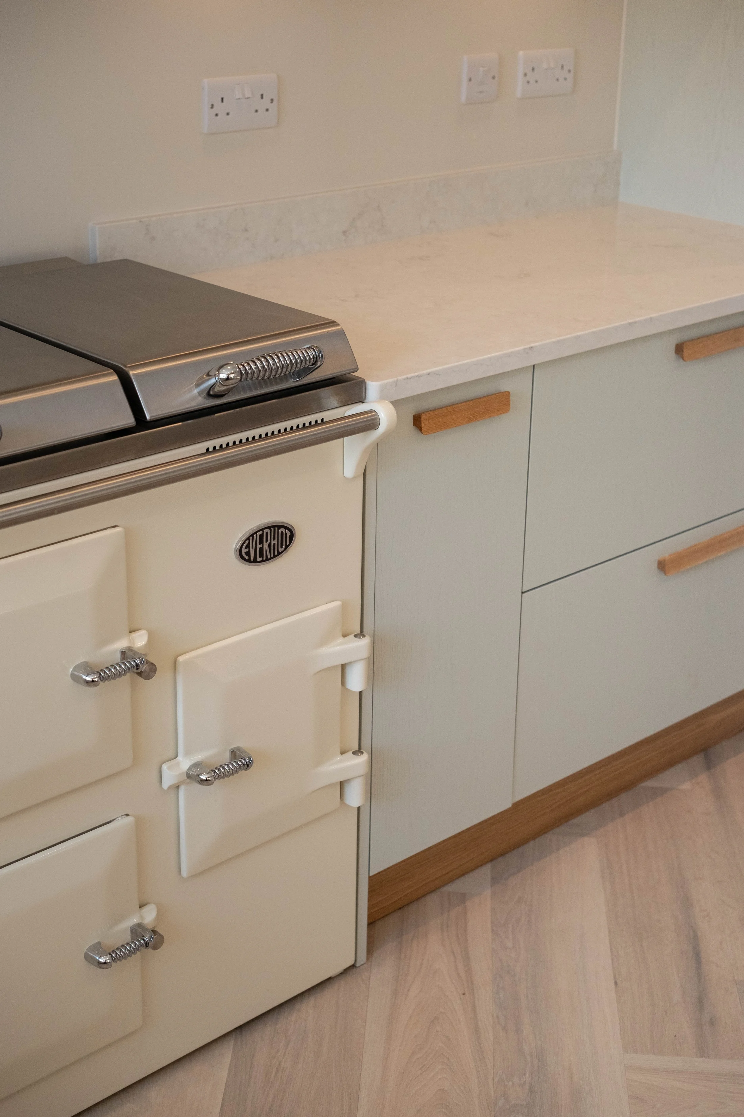 A cream-colored vintage-style gas stove with three small ovens and a metal coil above the burners is positioned next to white cabinetry with wooden handles, laminate countertops, and beige tile flooring.