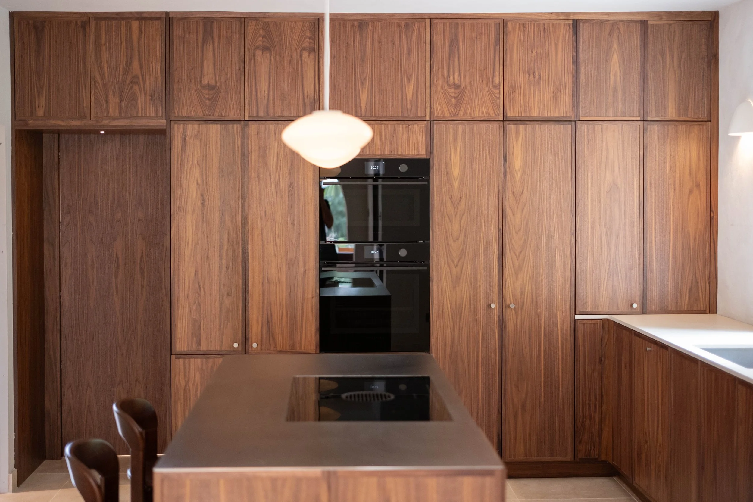 View of a kitchen with a large wooden cabinet wall, black built-in oven, and a beige countertop. A white hanging light fixture is visible in the foreground.