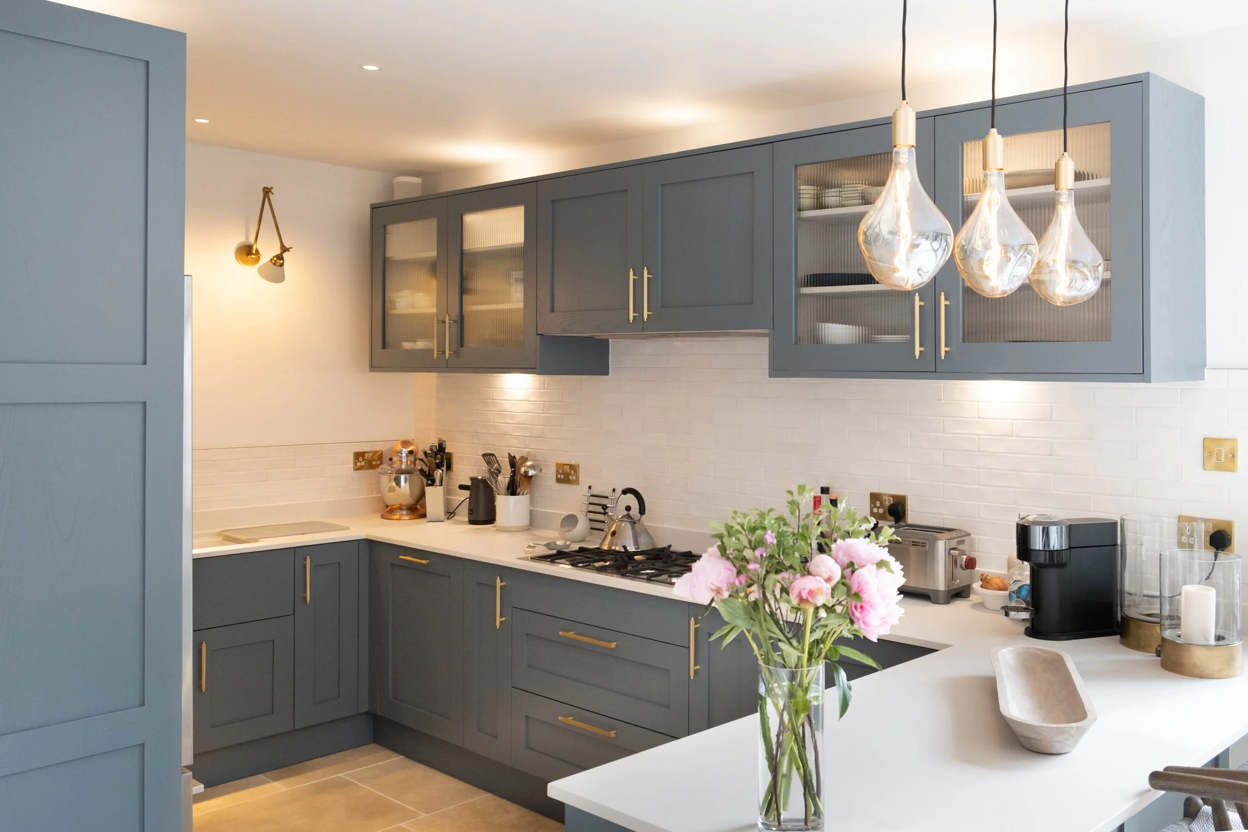 Modern kitchen with dark blue cabinets, gold handles, white countertops, and hanging glass pendant lights. A bouquet of pink flowers is on the counter, with various appliances and utensils visible.