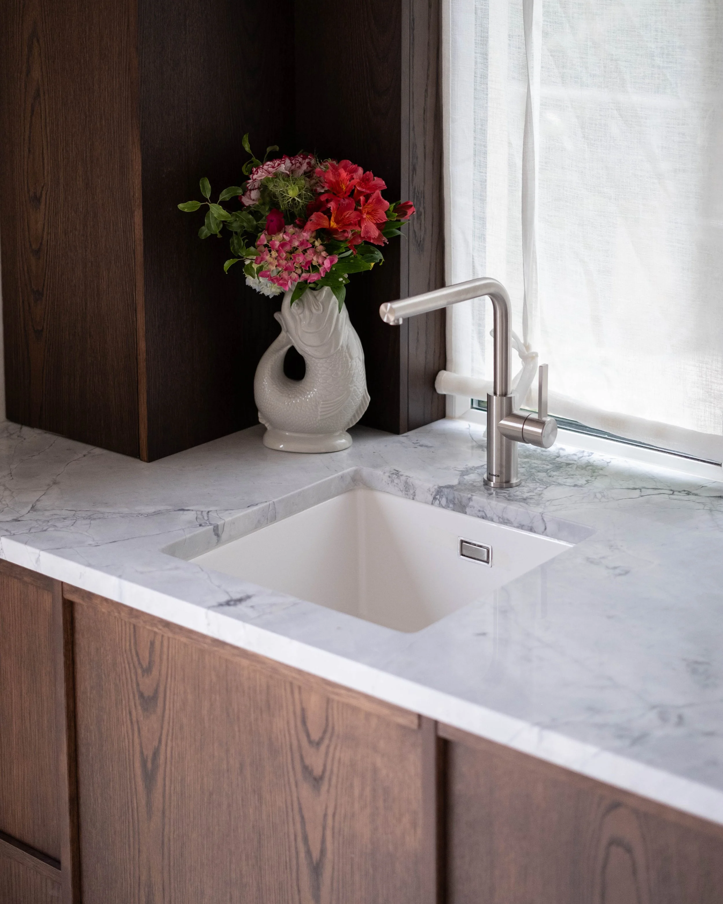 A kitchen countertop with a marble surface, a modern stainless steel faucet, a dark wooden cabinet, a decorative white ceramic vase shaped like a fish with pink and red flowers, and a white sheer curtain in the background.