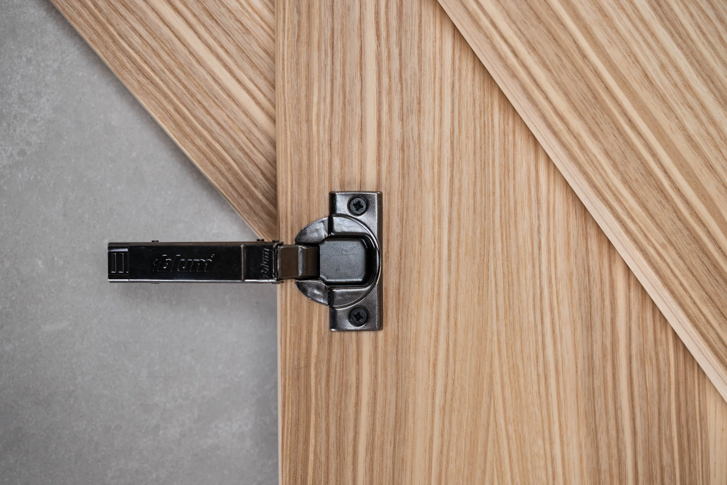 Close-up of a wood door with a metal hinge and a concealed door closer, set against a concrete wall.
