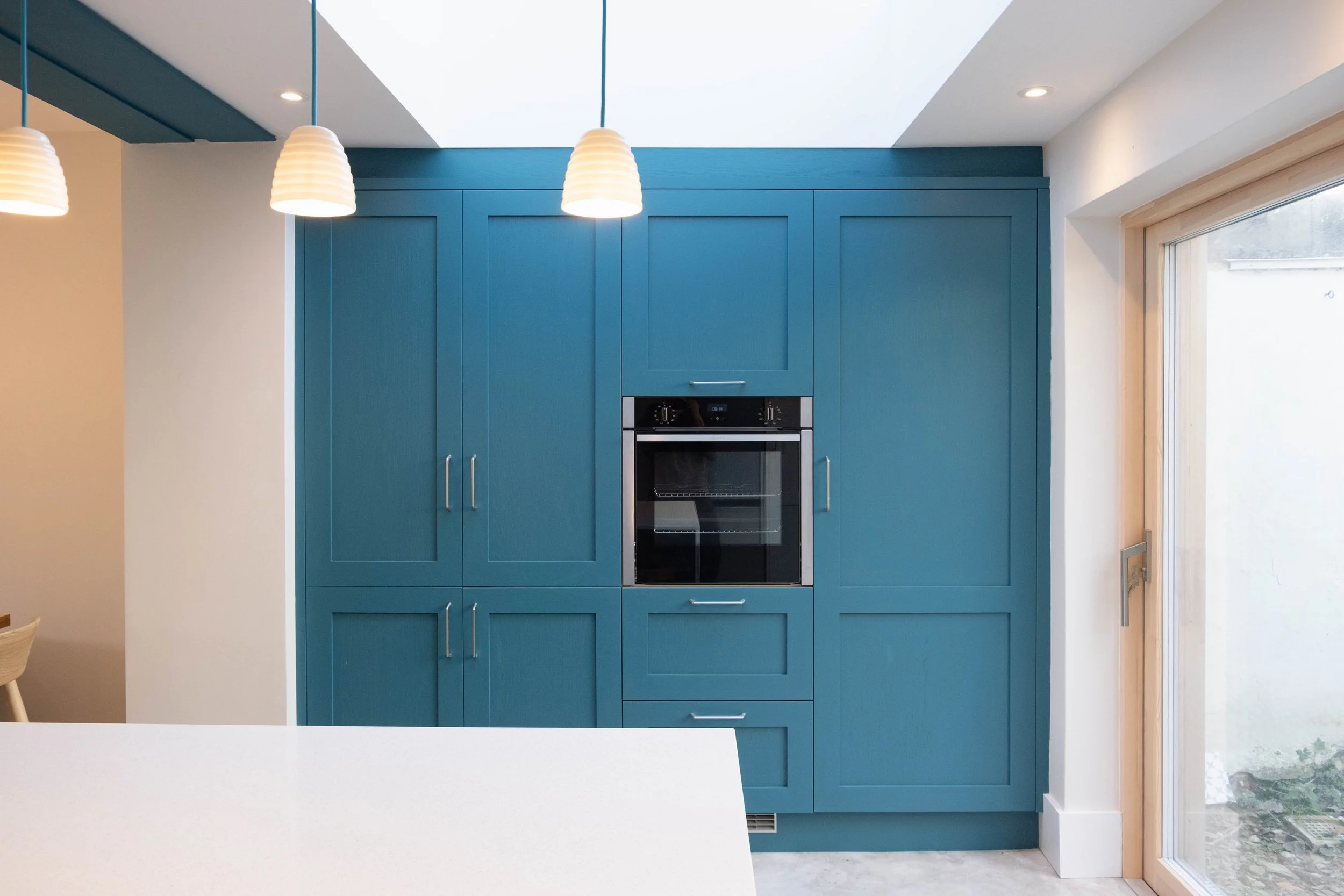 The image shows a modern kitchen with vibrant blue cabinets and built-in oven. There is a white countertop in the foreground and a sliding glass door on the right letting in natural light. Three white hanging pendant lights are suspended from the ceiling.