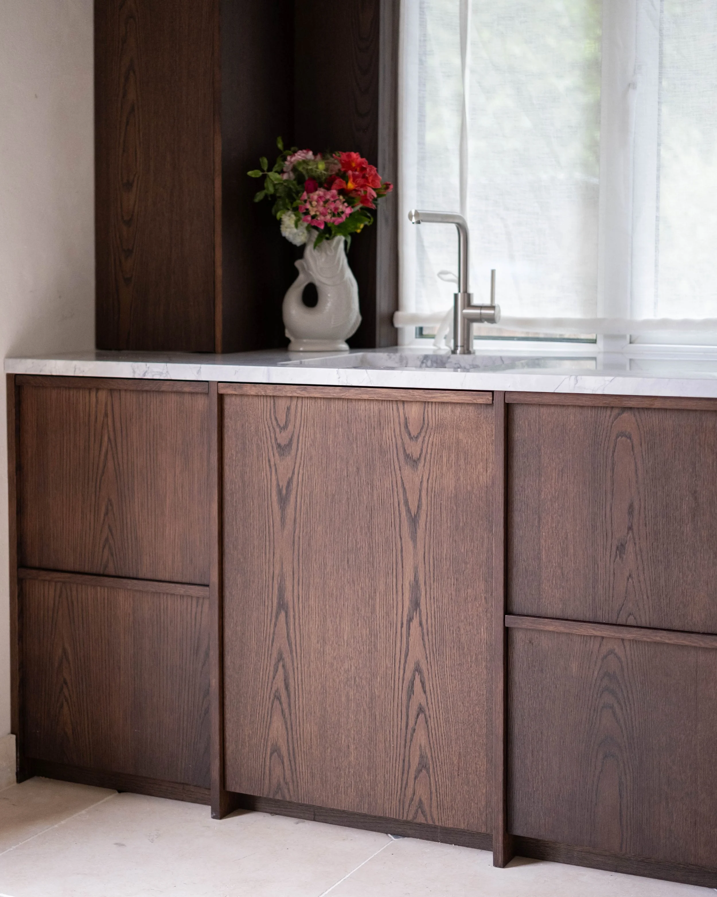 A kitchen sink area with dark wooden cabinets, a white marble countertop, a modern silver faucet, a window with sheer curtains, and a vase of pink and white flowers on the counter.