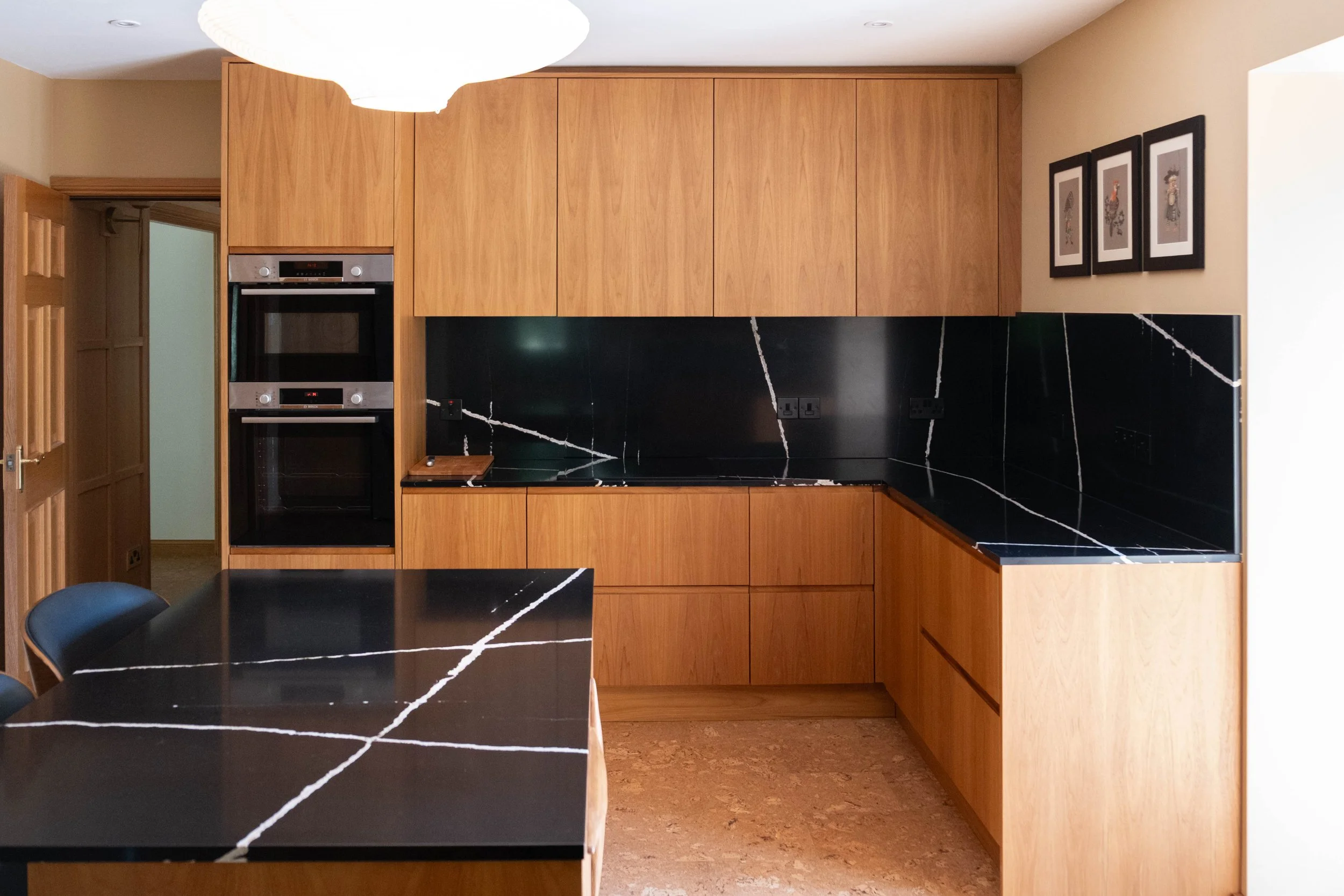 A modern kitchen with wooden cabinets, black marble countertops with white veining, and a built-in double oven. There is a black dining table with a chair, and three framed pictures of roosters on the wall. The floor is brown cork and there is a larg