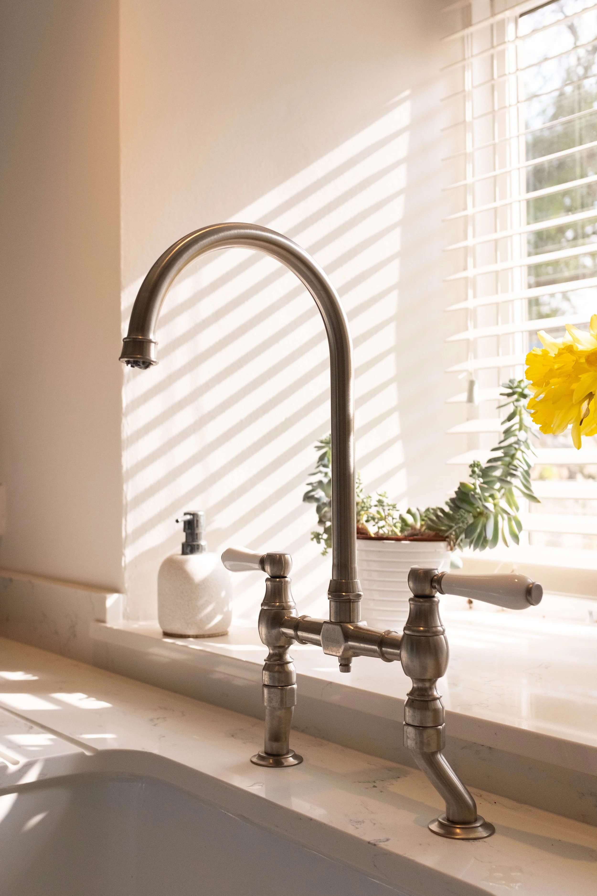 Close-up of a kitchen sink faucet with silver finish, set on a white marble countertop. Sunlight streams through window blinds, casting striped shadows on the wall behind. A white soap dispenser and a potted plant with yellow flowers are also visible