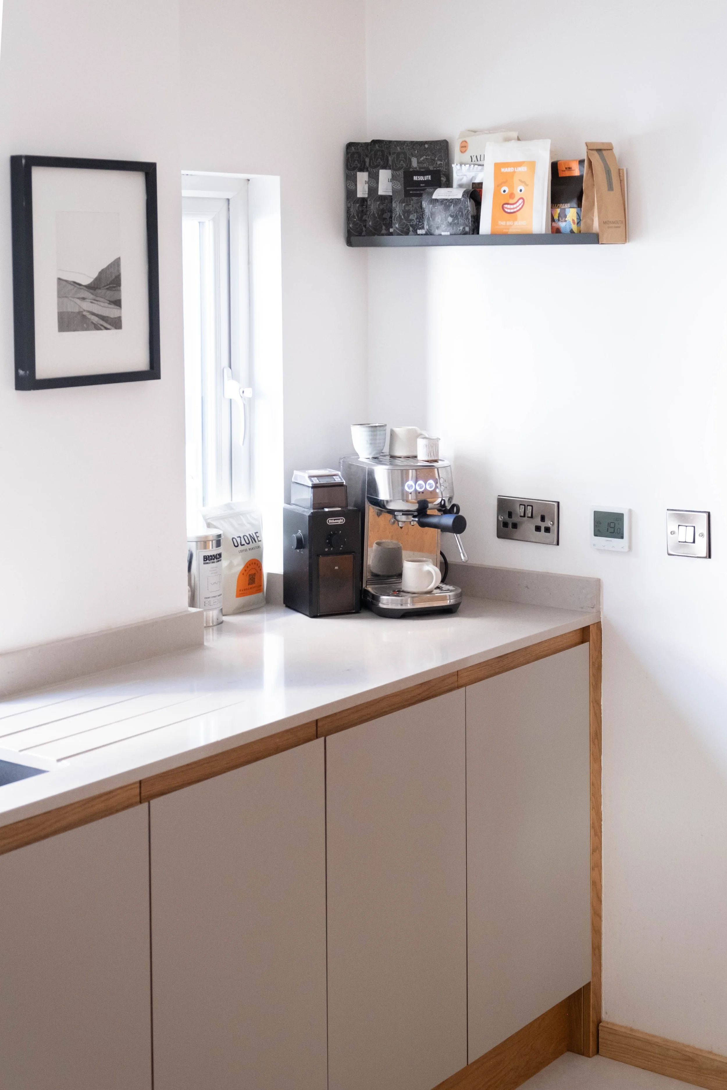 A kitchen corner with a white countertop, cream-colored cabinets with wooden trim, a coffee machine, a mug, and paper bags. A black picture frame with artwork hangs on the wall, and a window with natural light is visible. A black shelf holds books an